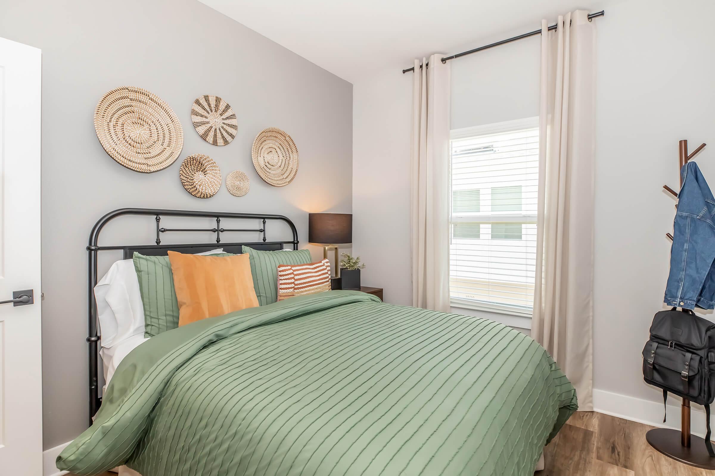 A cozy bedroom featuring a black metal bed frame with green and orange bedding. Decorative wall art made of woven materials is arranged above the bed. A lamp is placed on a nightstand, and soft curtains frame a window. A coat rack with a jacket and a backpack adds practical touches to the space.