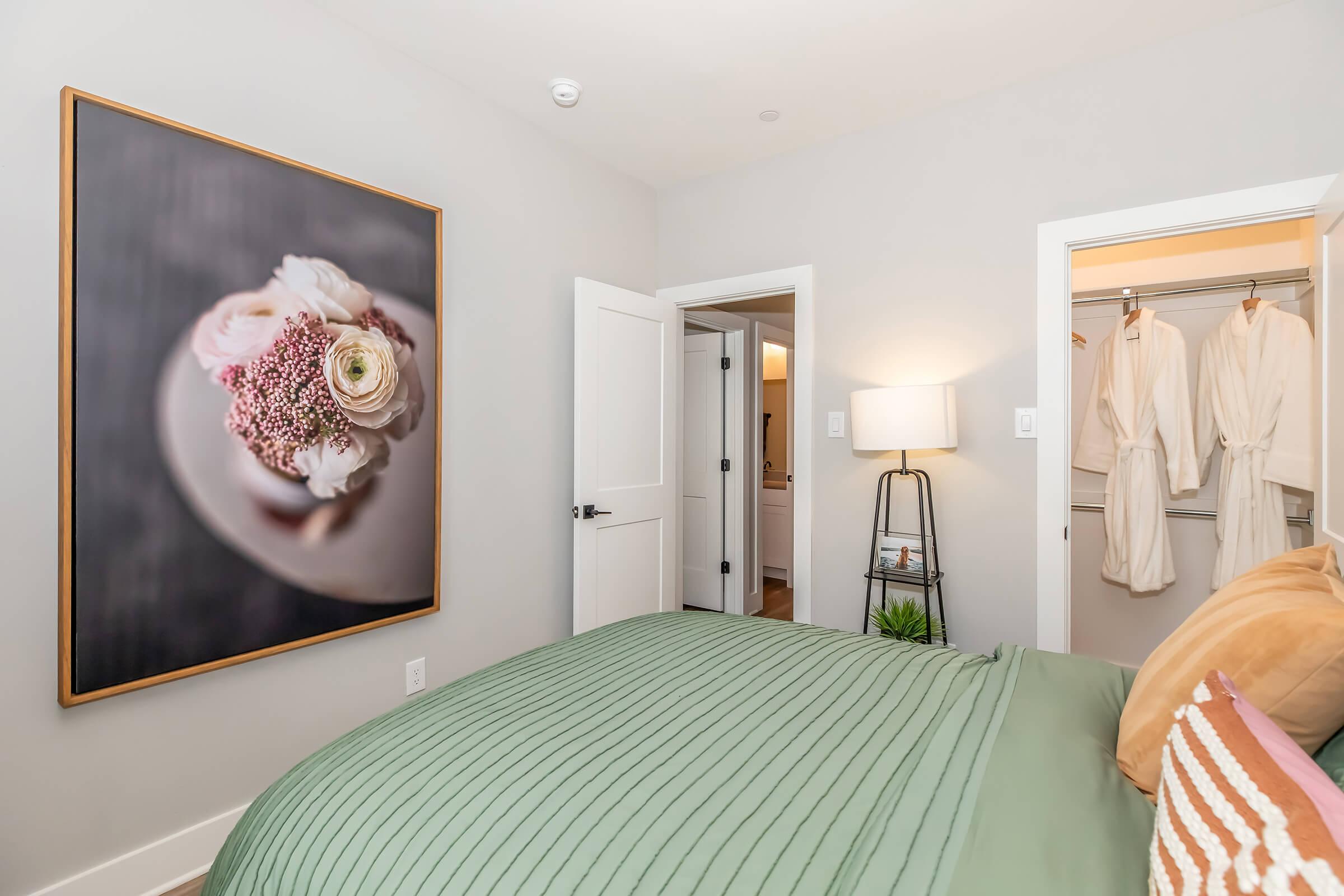 A cozy bedroom featuring a green striped duvet on a bed, a large floral artwork on the wall, a modern floor lamp, and two white bathrobes hanging in a closet. The room has soft neutral walls and a peaceful ambiance, with an open door leading to a bathroom area.