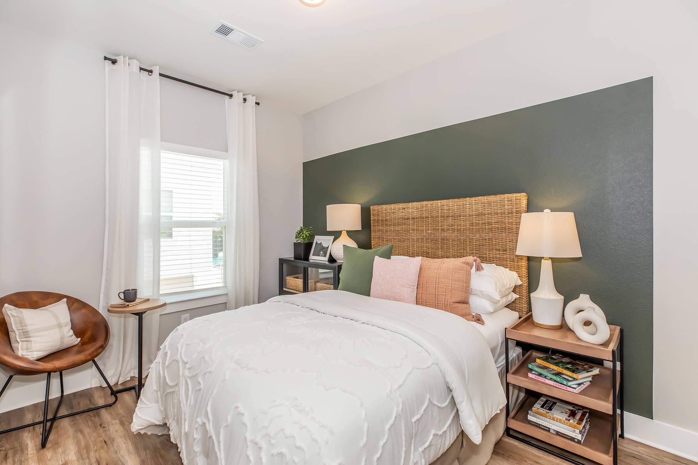 Cozy bedroom with a double bed featuring a white quilt and decorative pillows in shades of green, pink, and beige. A wooden headboard complements the dark green accent wall. A bedside table holds a lamp and books. Natural light enters through a window with sheer curtains, alongside a brown chair and small side table.