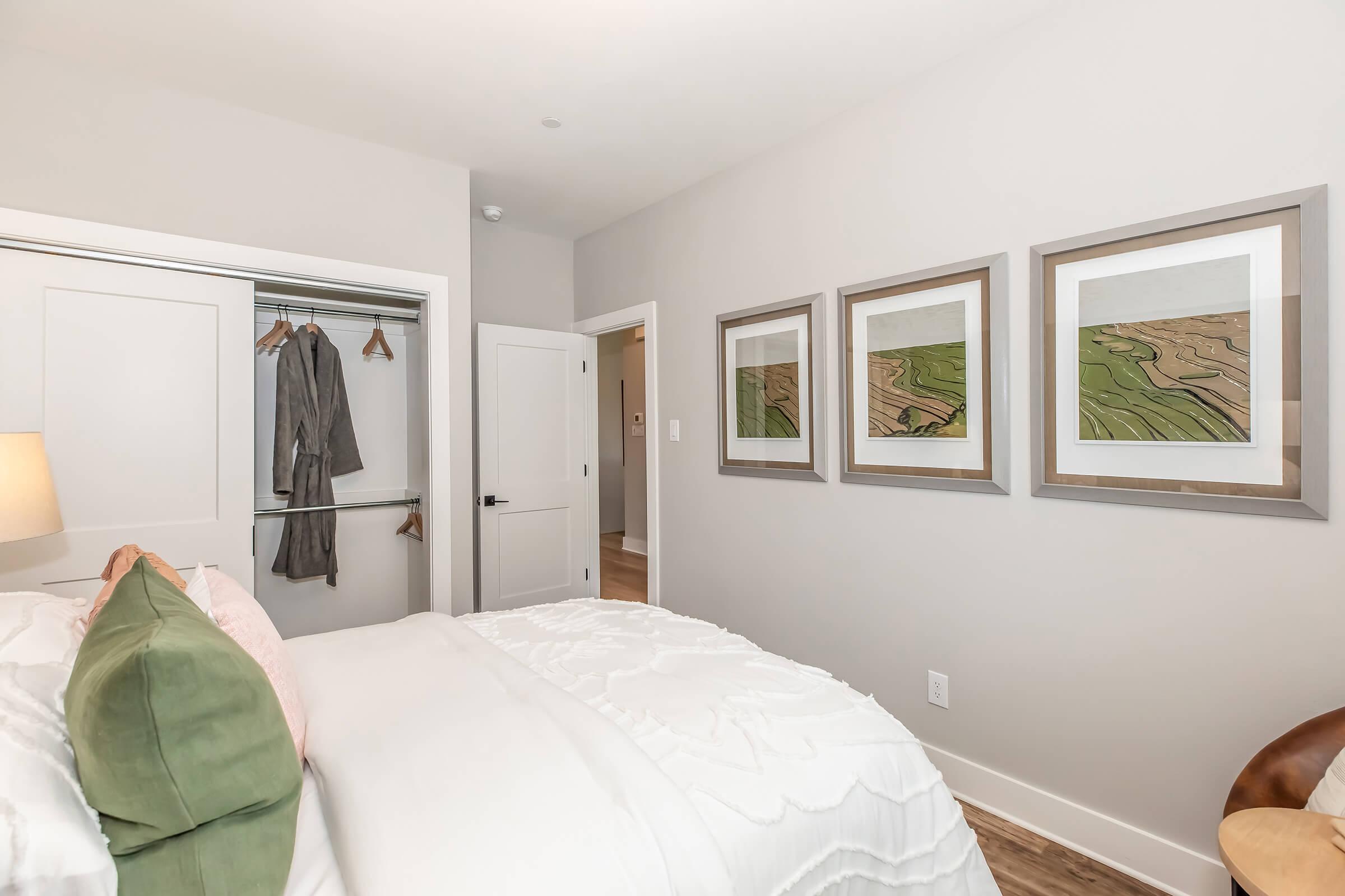 A modern bedroom featuring a neatly made bed with white bedding and green accent pillows. There is a wardrobe with clothing visible, and the walls are adorned with three framed artworks depicting abstract landscapes. The room has light gray walls and warm wooden flooring, creating a cozy atmosphere.