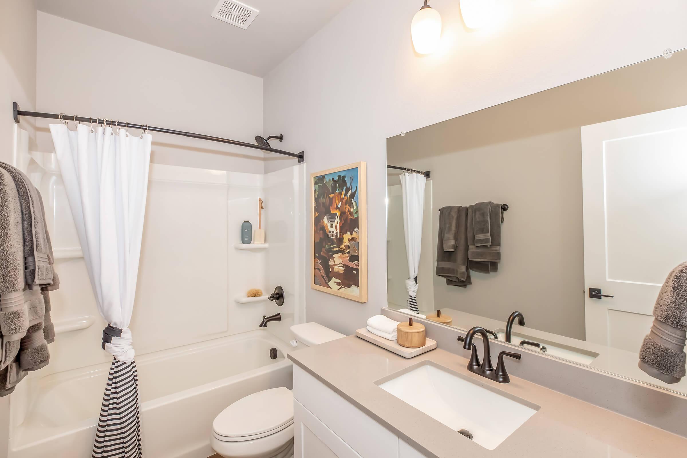 A modern bathroom featuring a bathtub with a shower, white curtains, and a sleek vanity with a rectangular sink. The room is adorned with a colorful wall art piece, neatly folded towels, and contemporary light fixtures. The color scheme is neutral, creating a clean and inviting atmosphere.