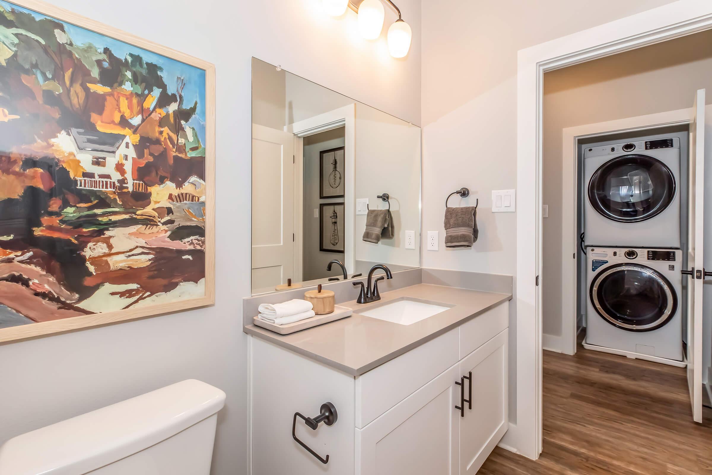 A modern bathroom featuring a double sink vanity with a mirror above, a colorful painting on the wall, and a doorway leading to a laundry area with stacked washer and dryer. The decor includes neutral tones and minimalistic elements, enhancing the bright and airy atmosphere.