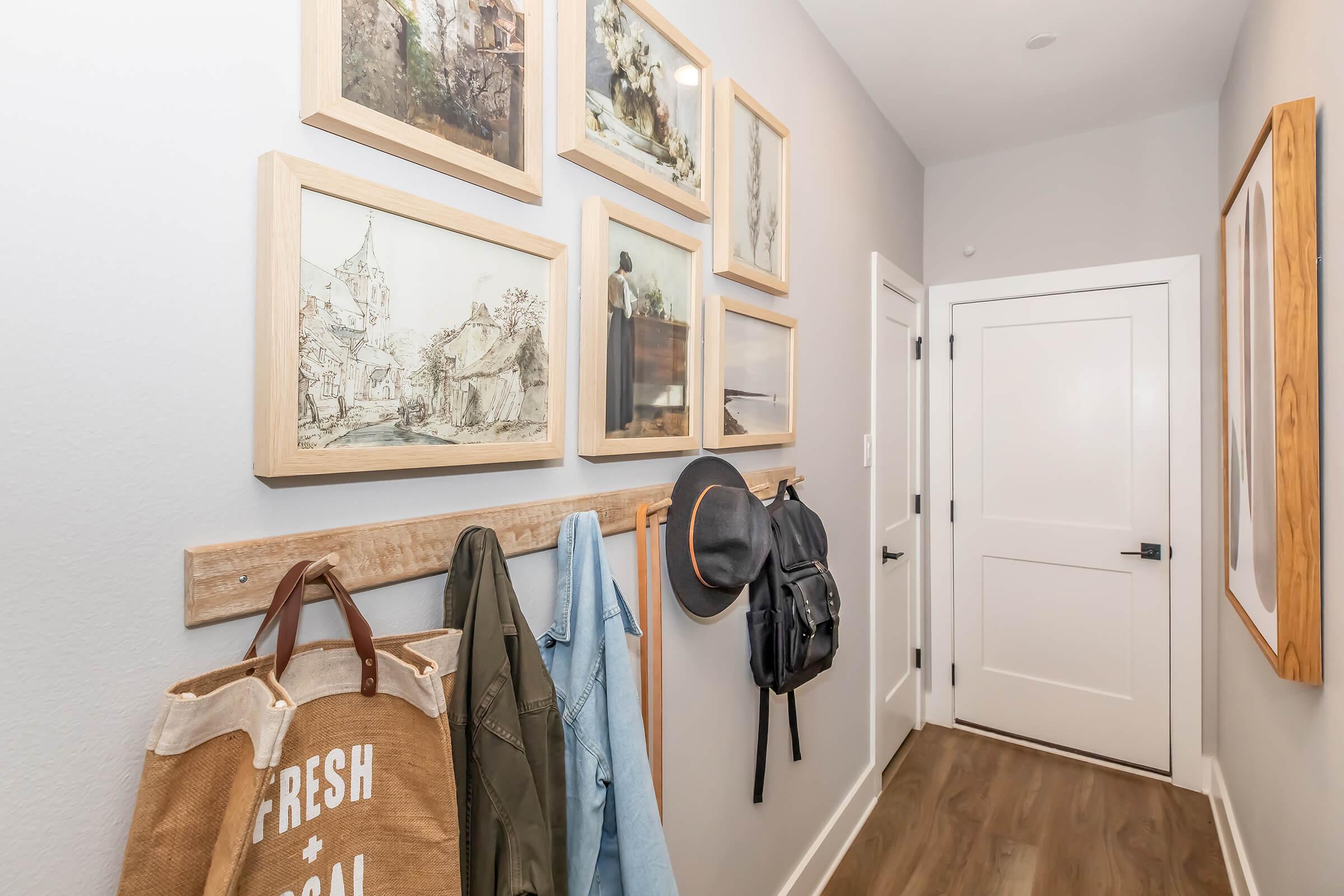 A modern hallway featuring light gray walls and wooden accents. There are several framed artworks hanging on the wall, showcasing various landscapes and figures. A coat rack holds a beige tote bag, a blue jacket, and a black backpack. A white door is visible at the end of the hallway.