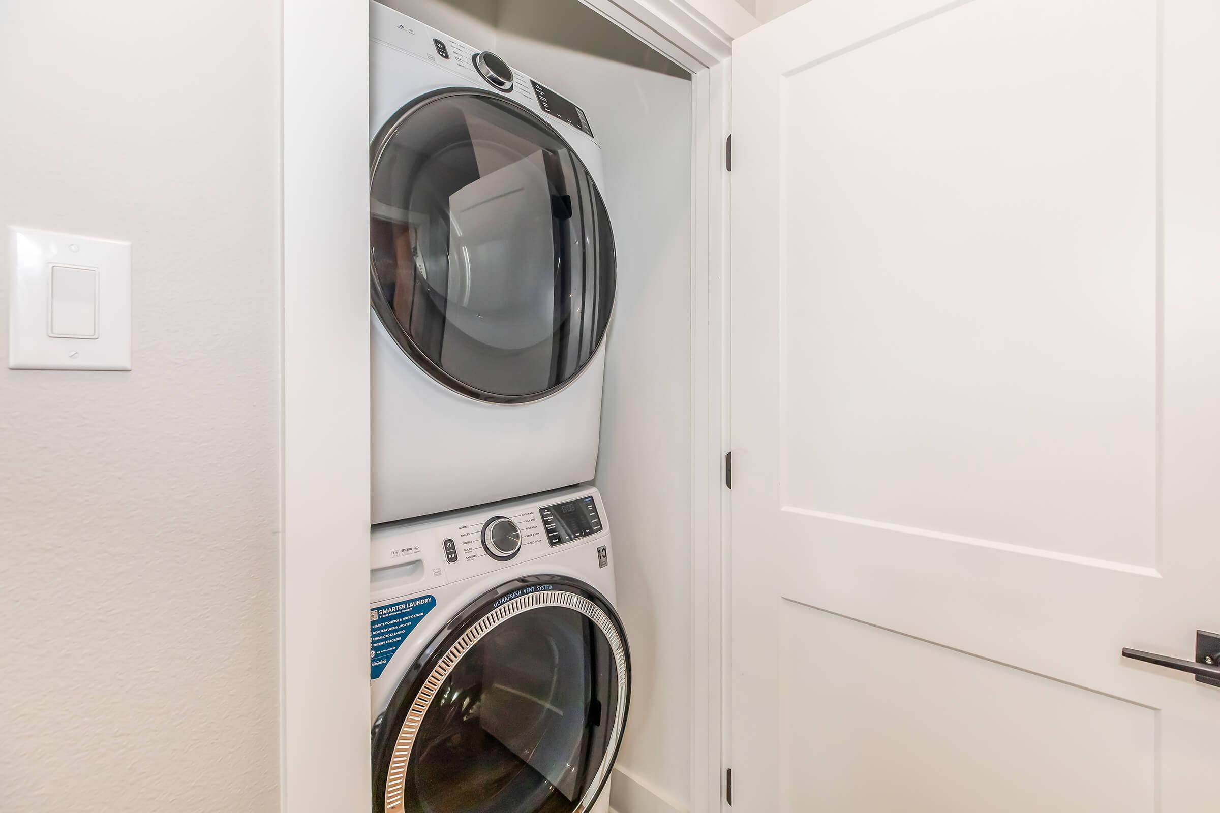 A stackable washer and dryer located in a small laundry closet, with a white door partially closed. The appliances have modern designs, with controls visible on the front of the dryer. The walls are painted in a neutral color, creating a tidy and functional laundry space.