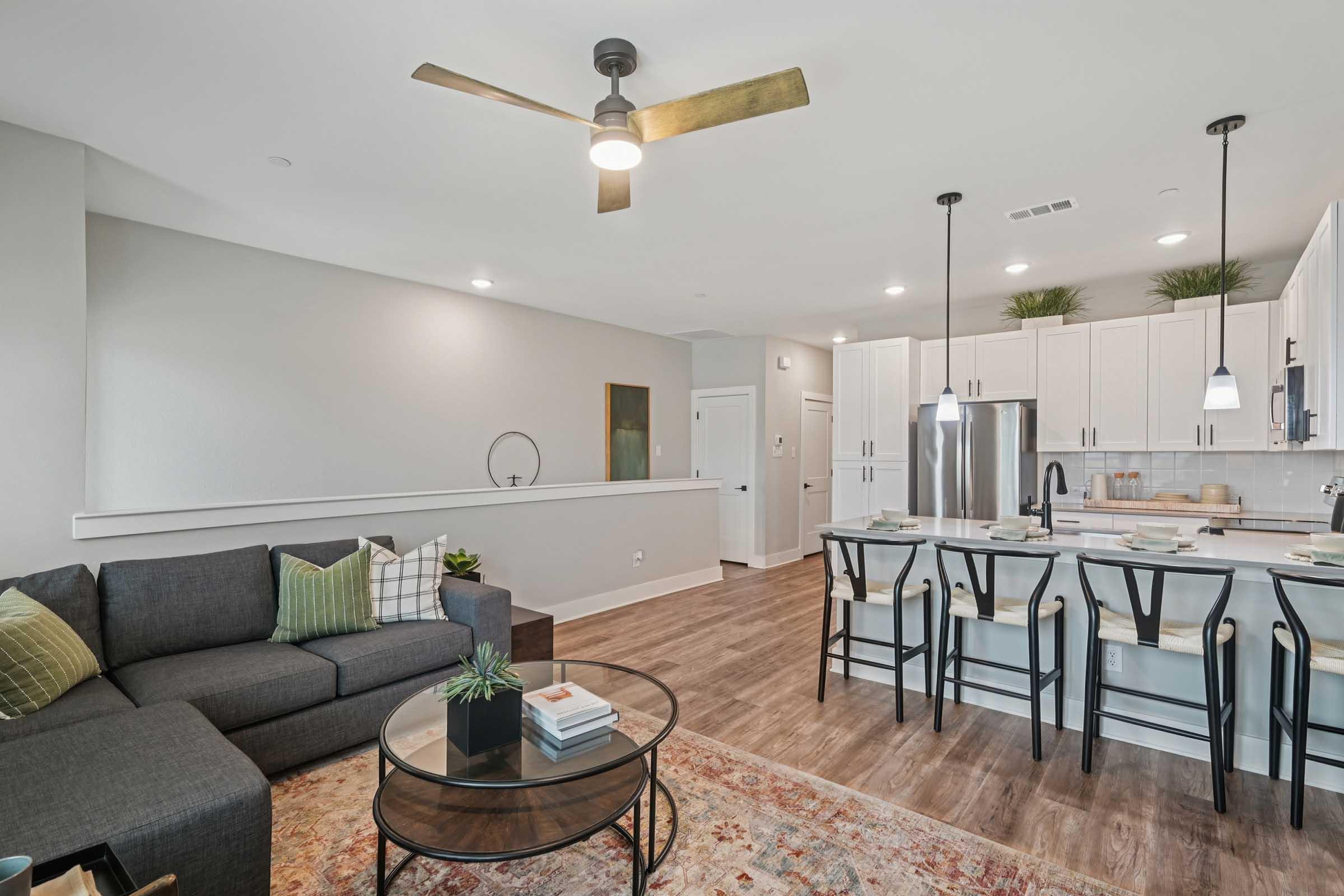 Modern living room and kitchen featuring a dark gray sectional sofa with green and white cushions, a round glass coffee table, and a stylish space with a ceiling fan. The kitchen has white cabinets, stainless steel appliances, and a breakfast bar with seating for four. A warm area rug adds comfort to the space.
