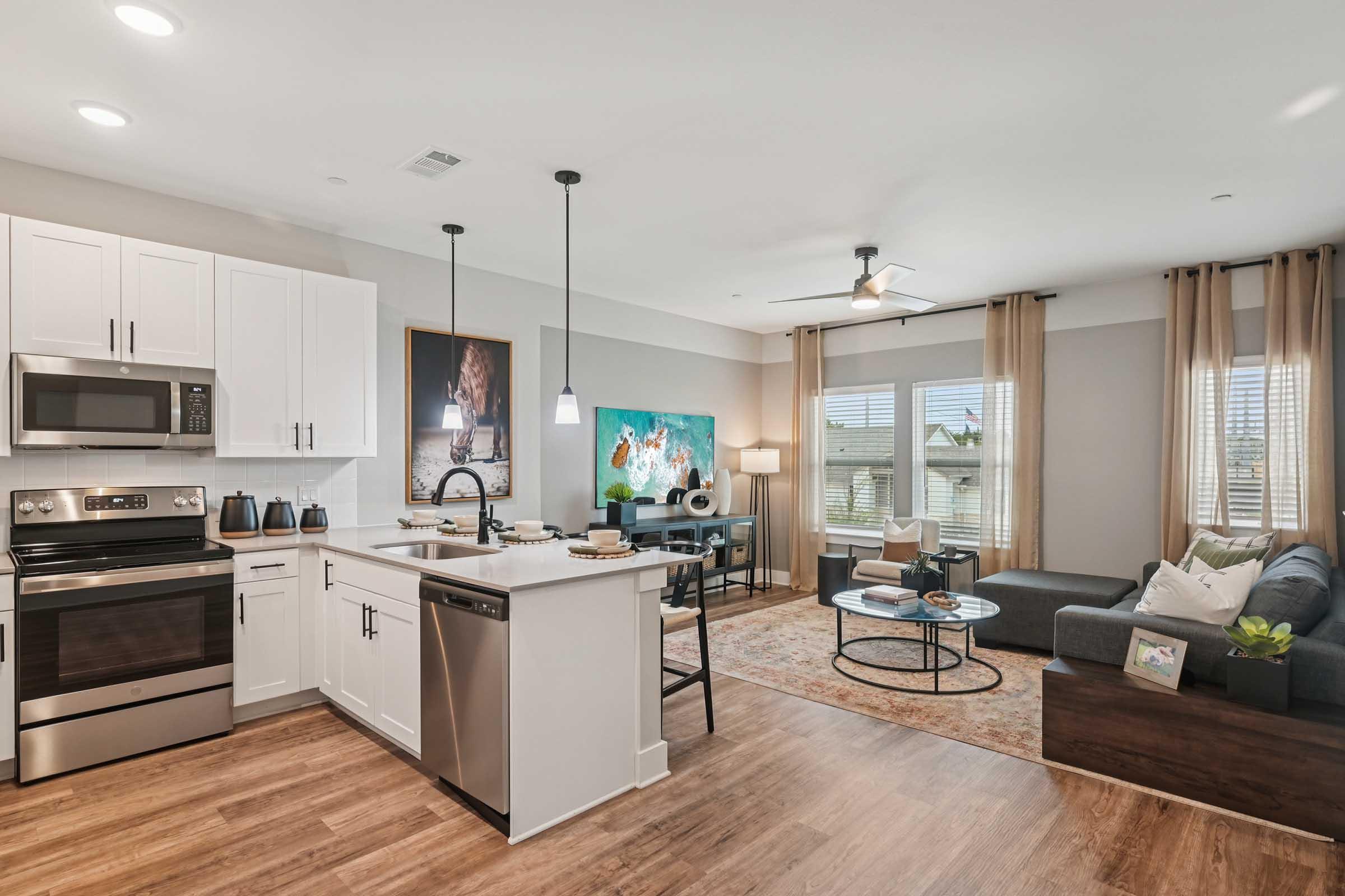 A modern kitchen and living area featuring white cabinets, stainless steel appliances, a dining table set, and a cozy sofa. Large windows with curtains allow natural light. The decor includes artwork and a colorful rug, creating a spacious and inviting atmosphere.