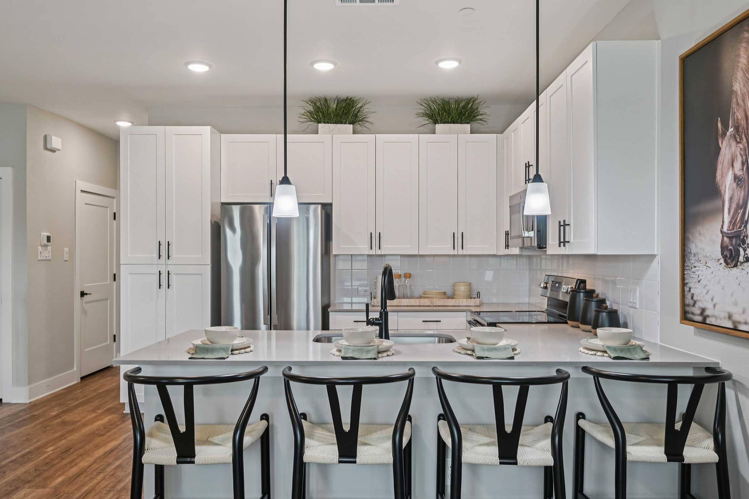 Modern kitchen with white cabinetry and granite countertops. Features a central island with four black chairs, pendant lighting, and stainless steel appliances. The backsplash is tiled, and there's a decorative horse painting on the wall. Lush greenery in the background adds a fresh touch.