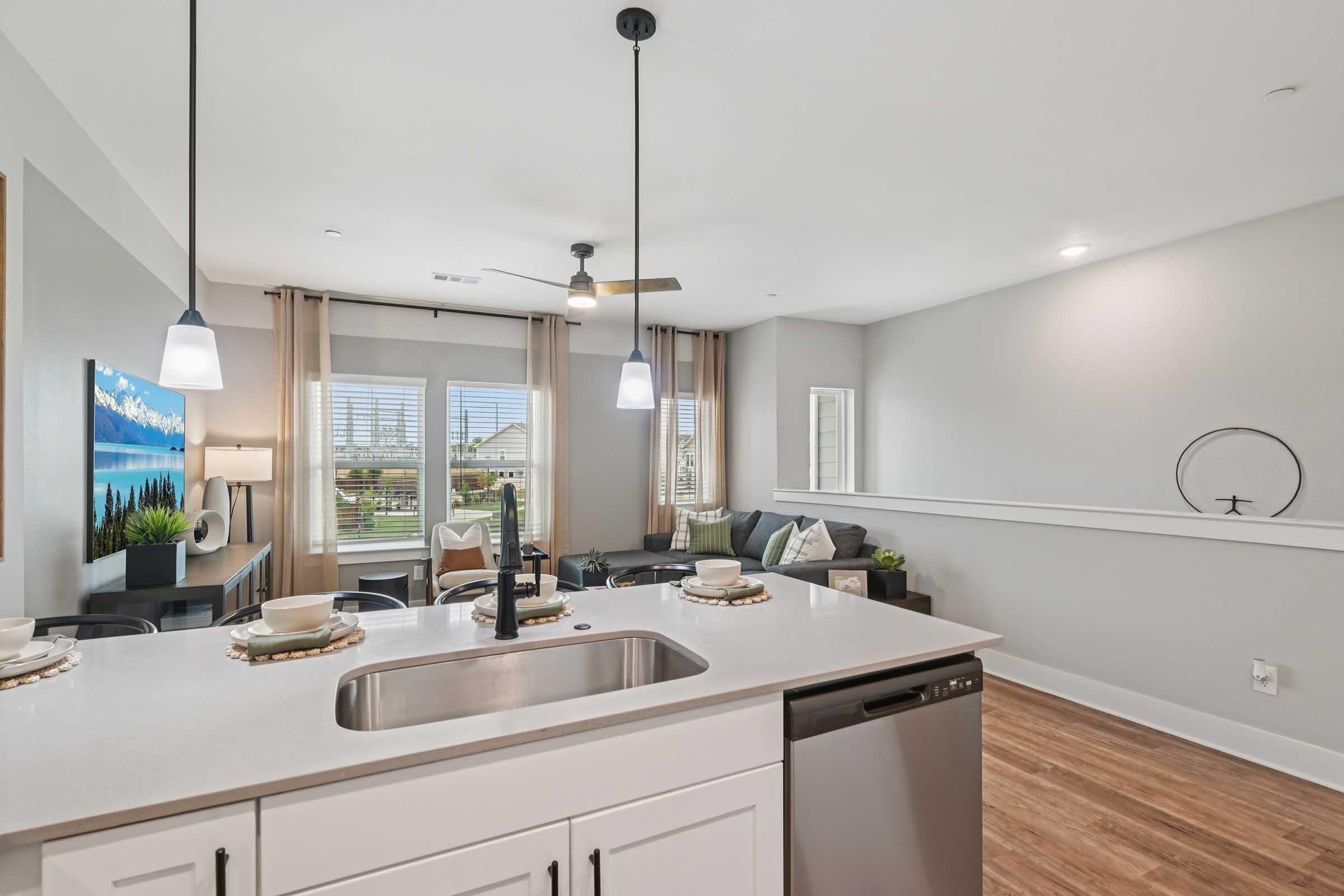 Modern kitchen view featuring a white countertop with a sink and stainless steel dishwasher. In the background, a living area with a comfortable seating arrangement, large windows allowing natural light, and decorative accents. Pendant lights hang above the kitchen island, creating a warm ambiance.