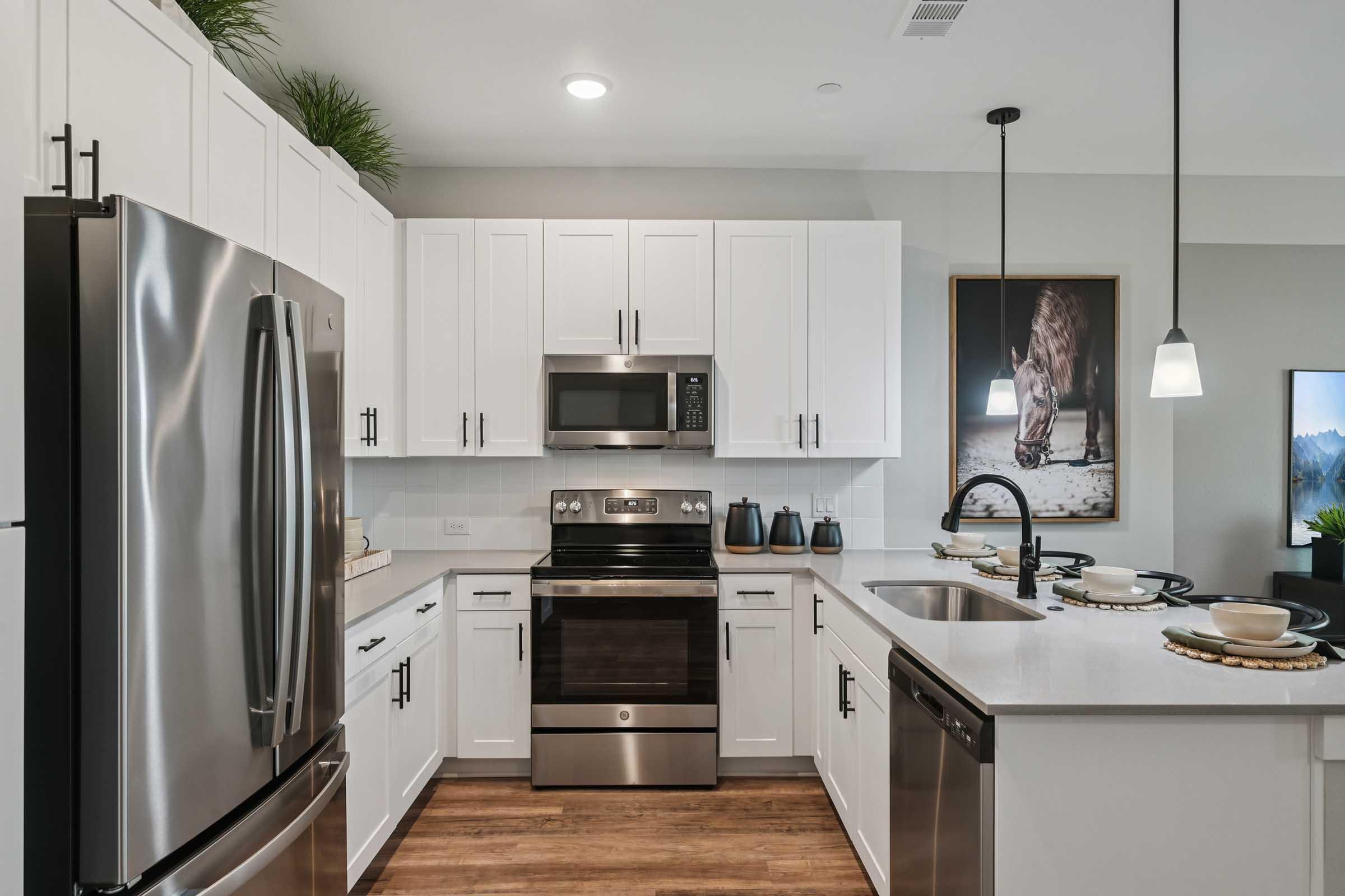 Modern kitchen interior featuring stainless steel appliances, white cabinets, and a light-colored countertop. A black faucet and sink are positioned in front of an island with dishware. A stylish art piece of a bear is hung on the wall, and pendant lights add a contemporary touch to the space.