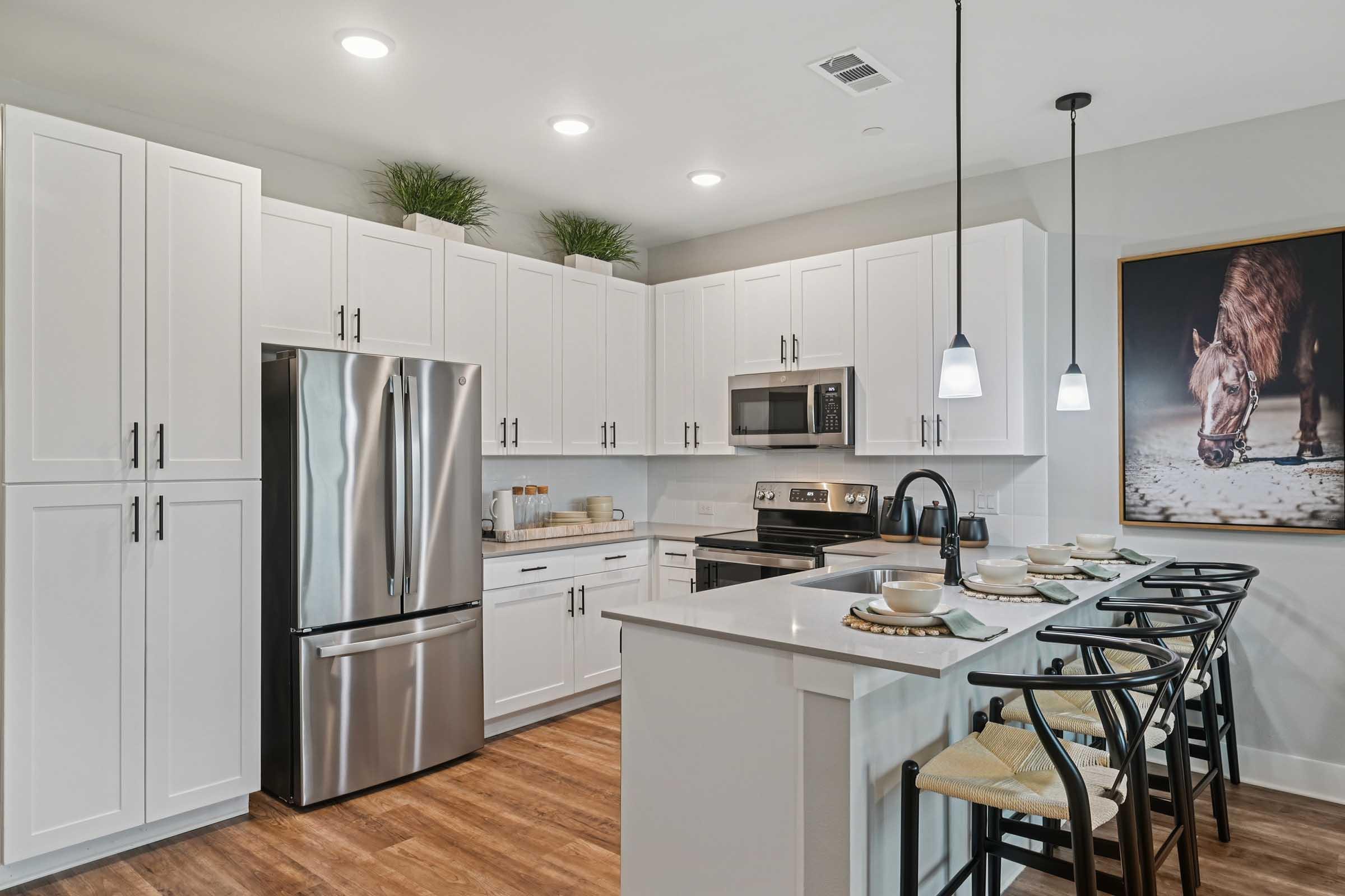Modern kitchen featuring white cabinetry, stainless steel appliances, and a large island with bar stools. The countertop is set for dining with plates and utensils. Soft lighting illuminates the space, complemented by decorative plants above the cabinets and an artwork featuring a horse on the wall.