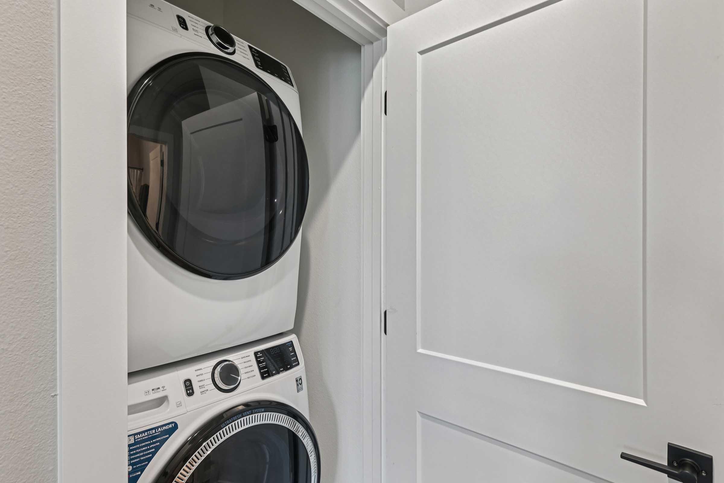 A stacked laundry unit consisting of a washer and dryer is positioned inside a small closet space. The closet door is partially open, revealing the white appliances against a light-colored wall. The space is minimal and organized, with a sleek design.