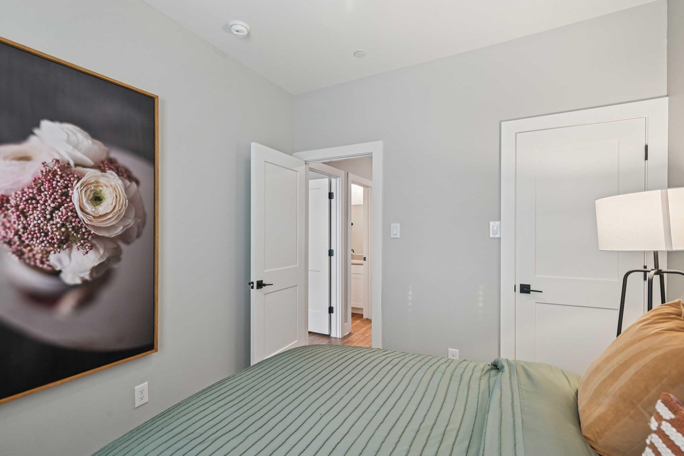 A cozy bedroom featuring a large floral artwork on the wall, a neatly made bed with a green cover, and soft lighting from a nearby lamp. The room has light-colored walls and an open door leading to a bathroom area, adding a sense of spaciousness and inviting ambiance.