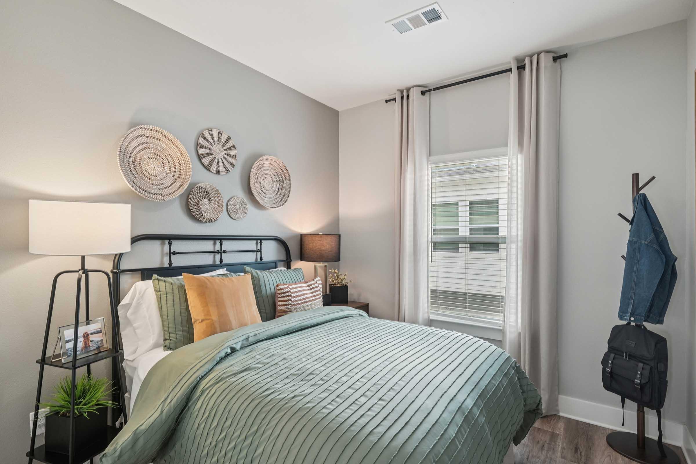 Cozy bedroom featuring a bed with a green textured comforter, decorative pillows, and a stylish headboard. The walls are adorned with woven wall art, and there is a lamp on a nightstand. A window with light curtains provides natural light, while a coat rack and potted plant add to the decor.