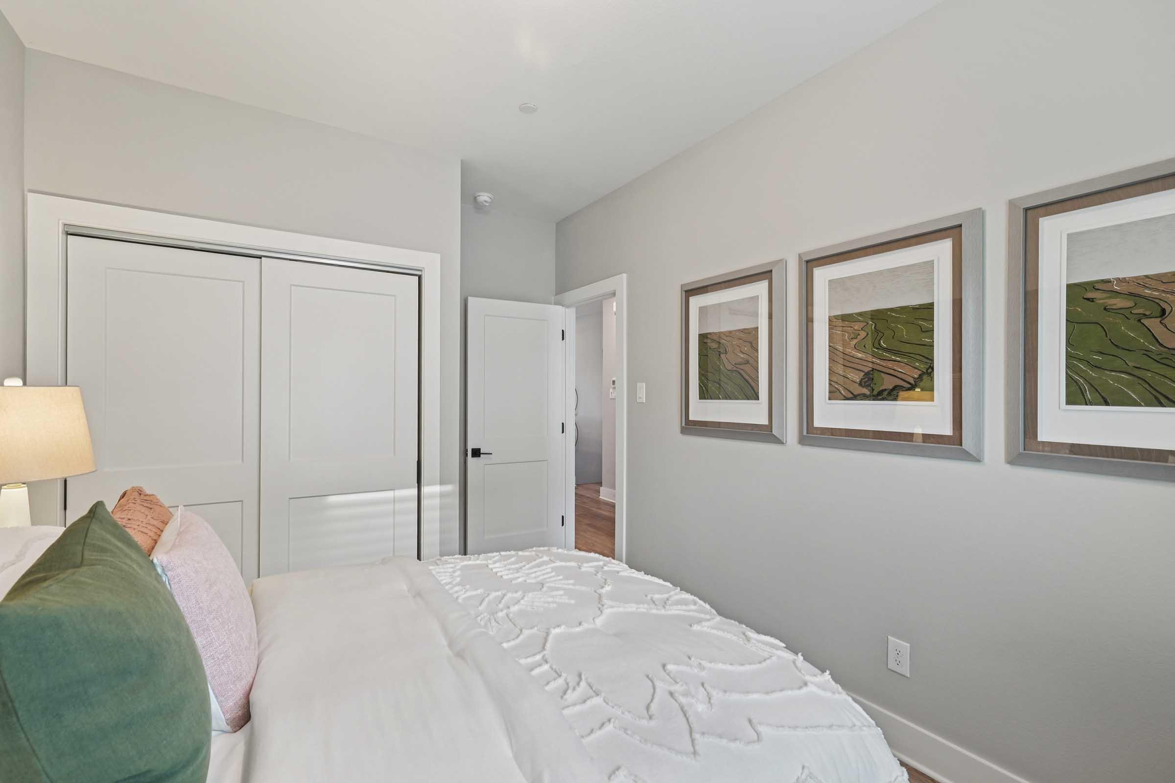A cozy bedroom featuring a neatly made bed with decorative pillows, soft lighting from a bedside lamp, and framed artwork on the wall. The room has light gray walls, a closet with sliding doors, and wooden flooring, creating a calm and contemporary atmosphere.