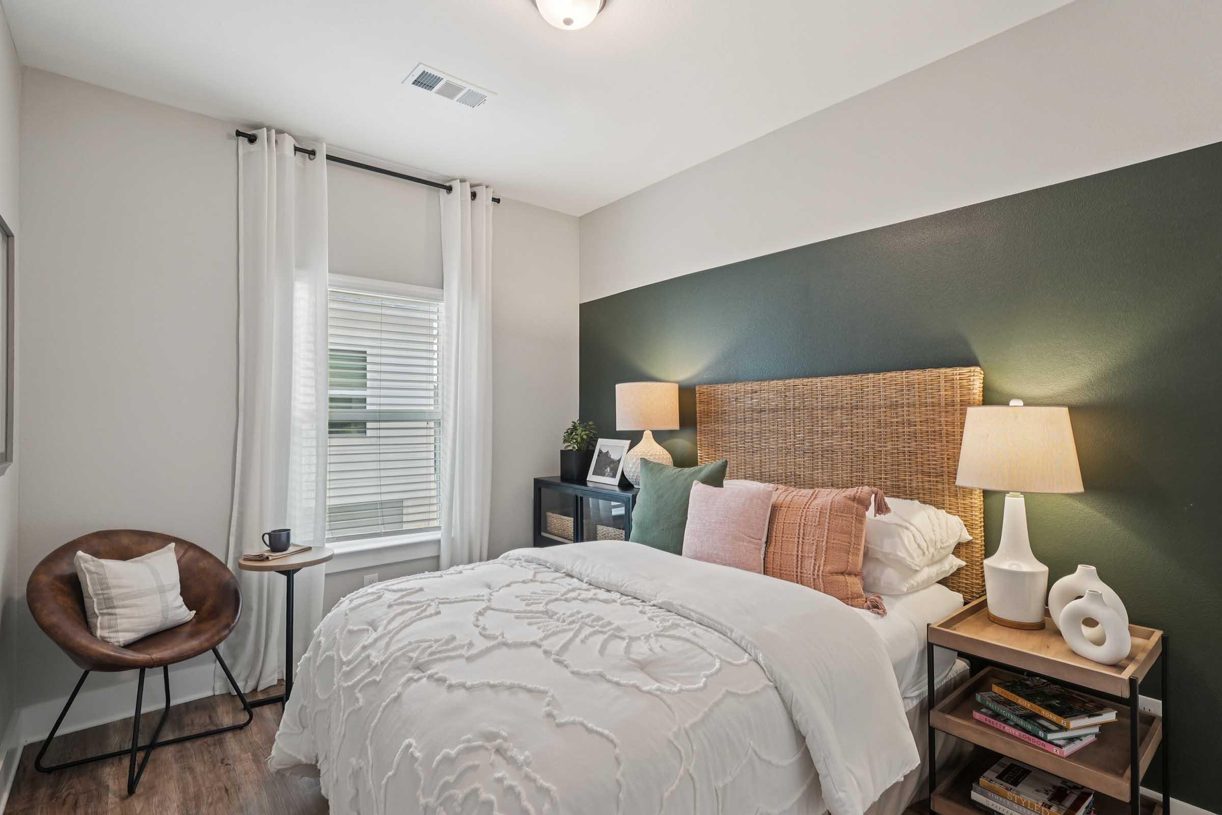 A cozy bedroom featuring a bed with a white decorative blanket and various pillows, a wooden headboard, and two bedside lamps. A chair and small table are positioned beside the bed. The walls are painted in a dark green accent, while natural light enters through a window covered with light curtains.
