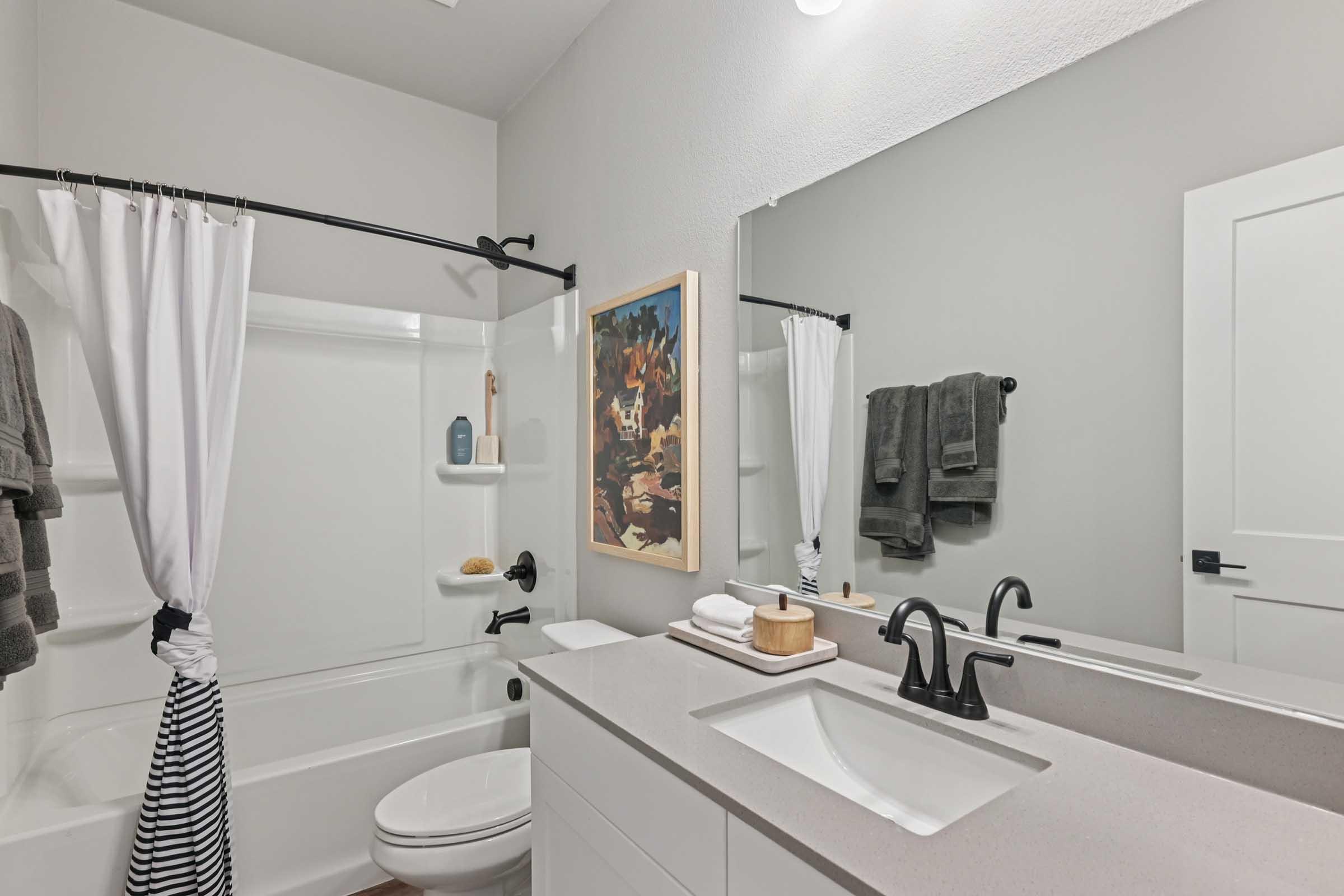 A modern bathroom featuring a white bathtub with a black shower curtain, a vanity with a sink, and a large mirror. The room is decorated with gray walls, towels hanging on hooks, and a piece of artwork on the wall. Natural light illuminates the space, creating a clean and inviting atmosphere.