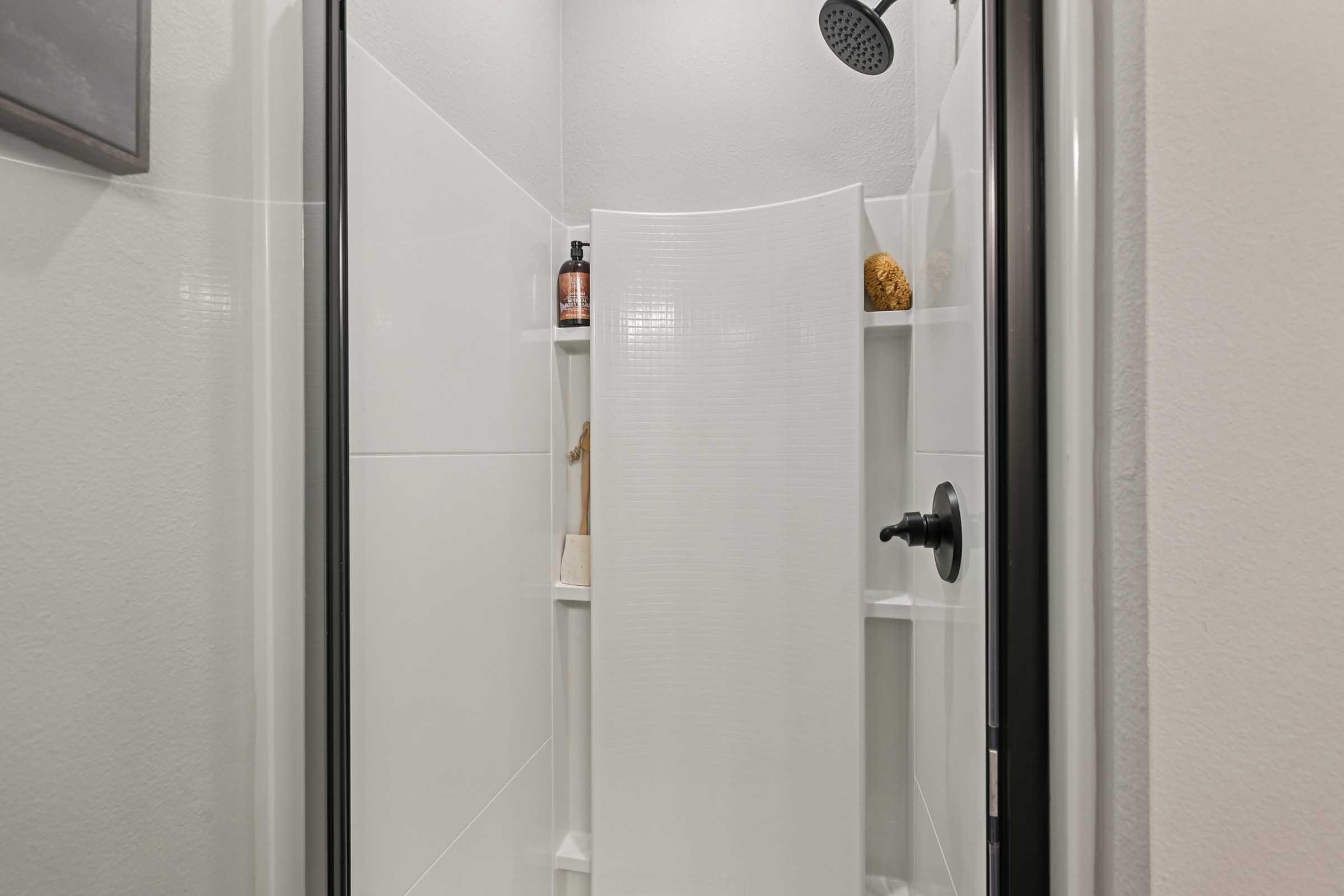 A modern shower stall featuring white tiled walls, a sleek black showerhead, and minimalist shelving stocked with toiletries. The design is clean and contemporary, emphasizing an open and inviting space.