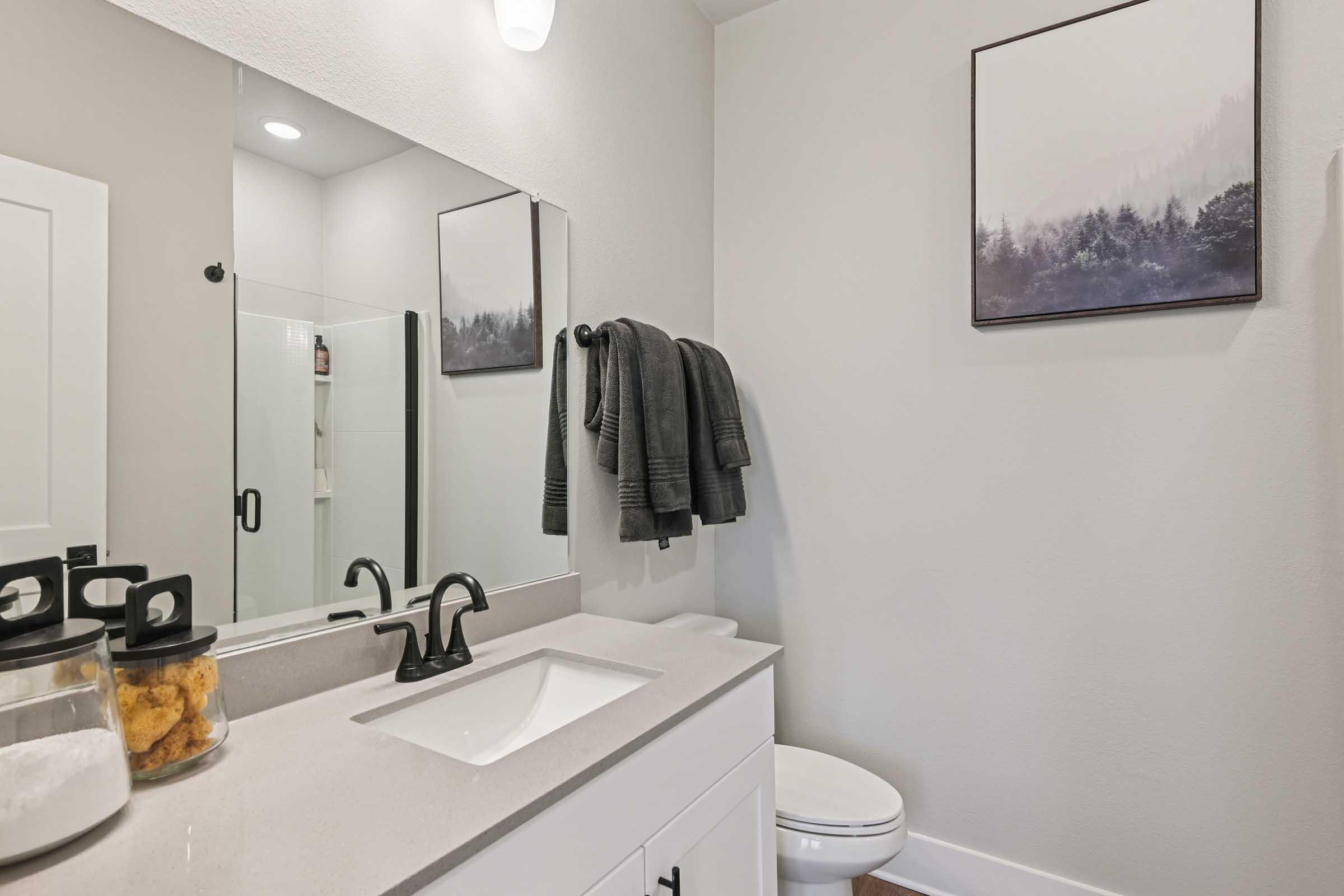 A modern bathroom featuring a white vanity with a sink, a mirror above it, and a shower area with glass doors. Two dark gray towels hang from a rack. On the countertop, there is a jar with golden bath salts. A framed artwork with a misty landscape hangs on the wall, complementing the neutral tones of the decor.