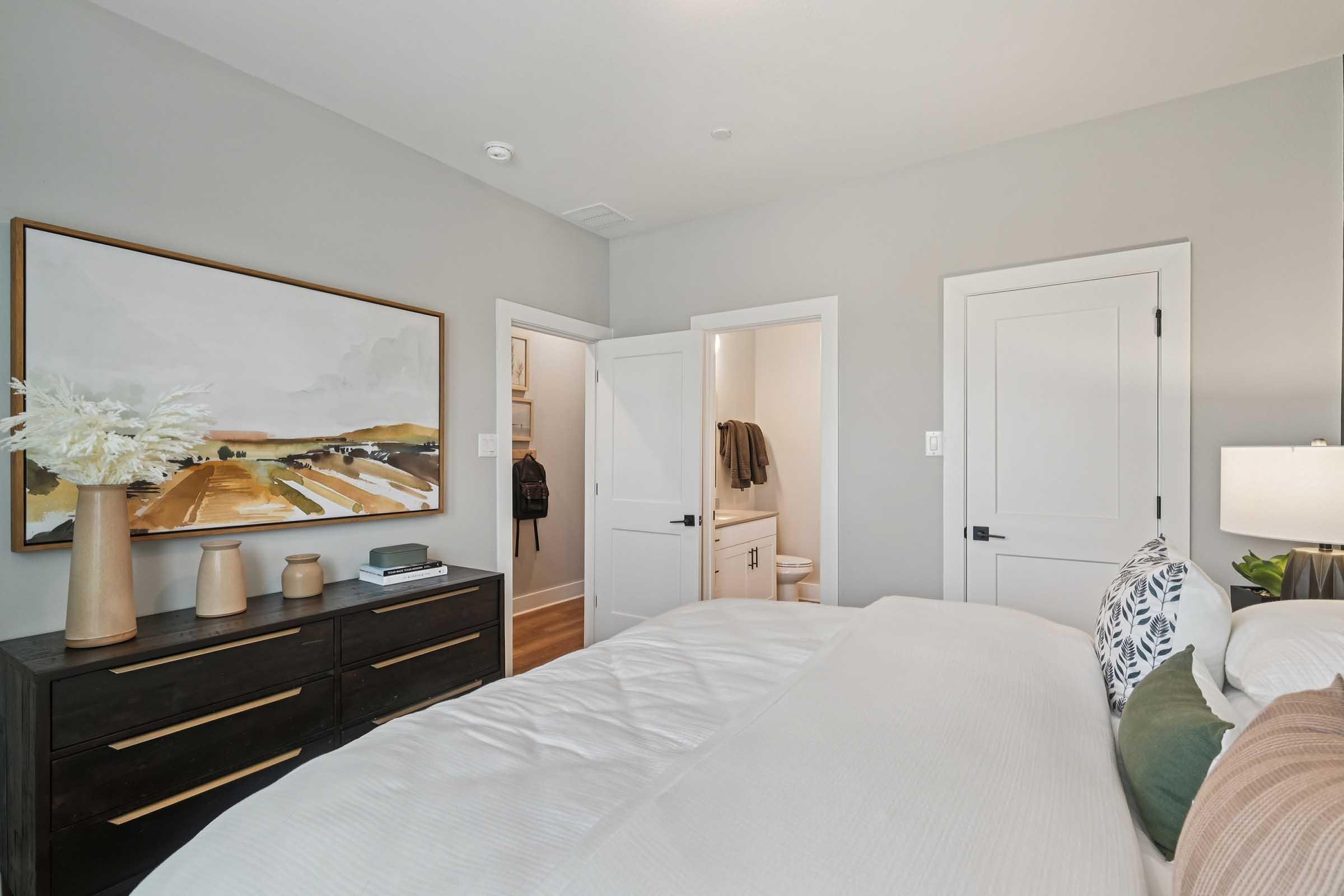 A modern bedroom featuring a large painting on the wall above a dark wood dresser. A white bed with decorative pillows is in the foreground. A door leads to a bathroom, and another door is slightly ajar, revealing a hallway. Soft, neutral colors dominate the decor, creating a cozy atmosphere.