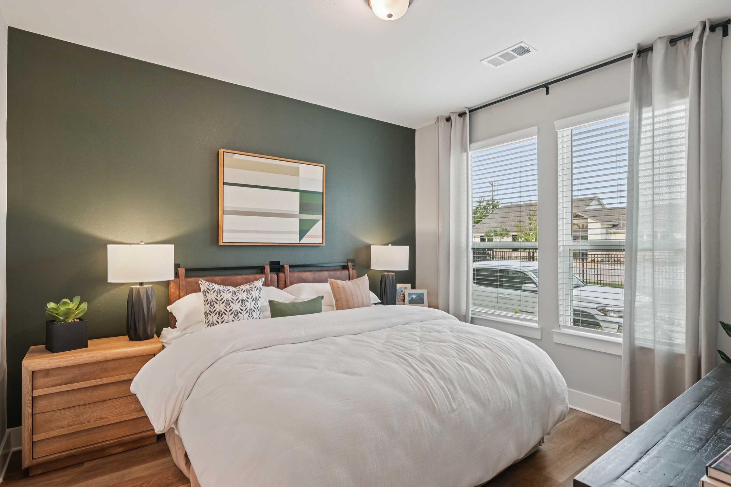 Cozy bedroom featuring a large bed with white bedding, wooden nightstands with table lamps, and a green accent wall. There are large windows with sheer curtains letting in natural light, and a modern piece of art hangs above the bed. A small plant adds a touch of greenery to the room.