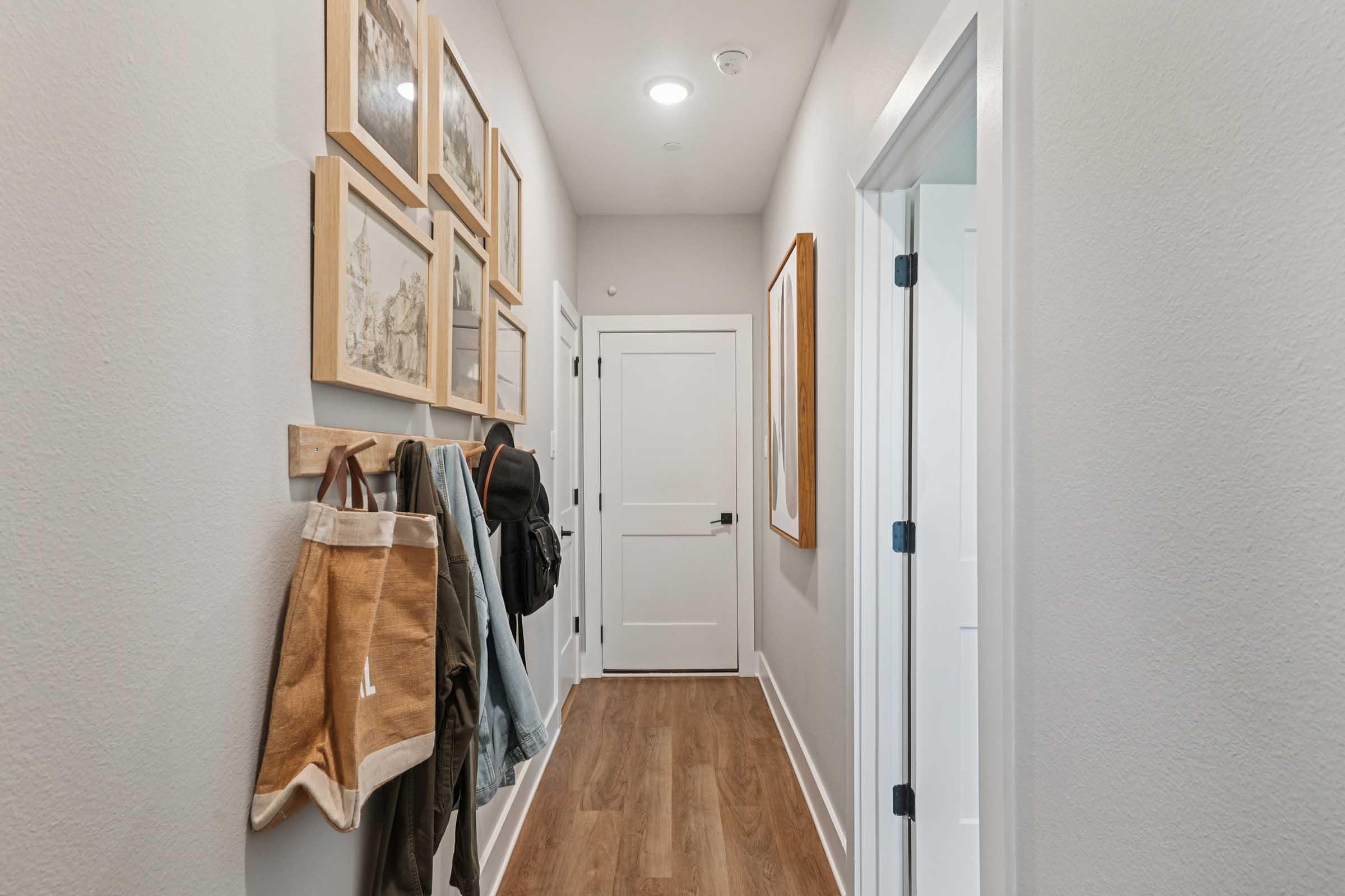 A narrow hallway featuring light gray walls and wooden flooring. The walls are adorned with framed artwork, and there are hooks with jackets and a backpack hanging. A closed door is visible at the end of the hallway, along with a partially open door leading to another room. The lighting is bright and inviting.
