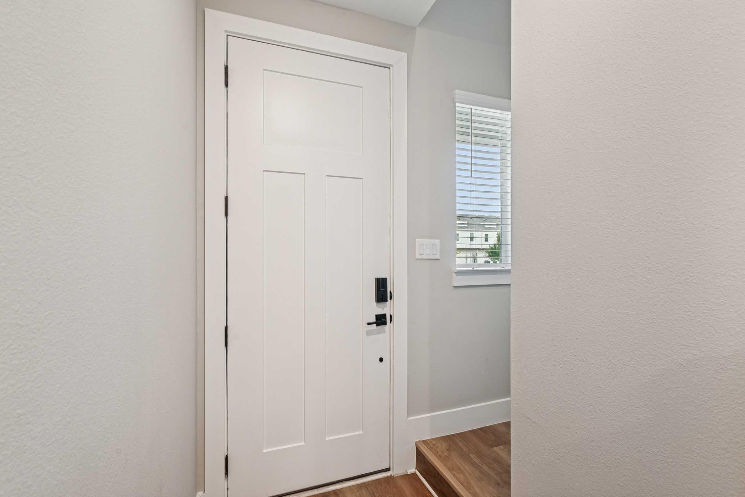 A clean, modern entryway featuring a white front door with black hardware, set within a light gray wall. To the side, a small set of stairs leads up to a bright space, complemented by a window with horizontal blinds allowing natural light to enter.