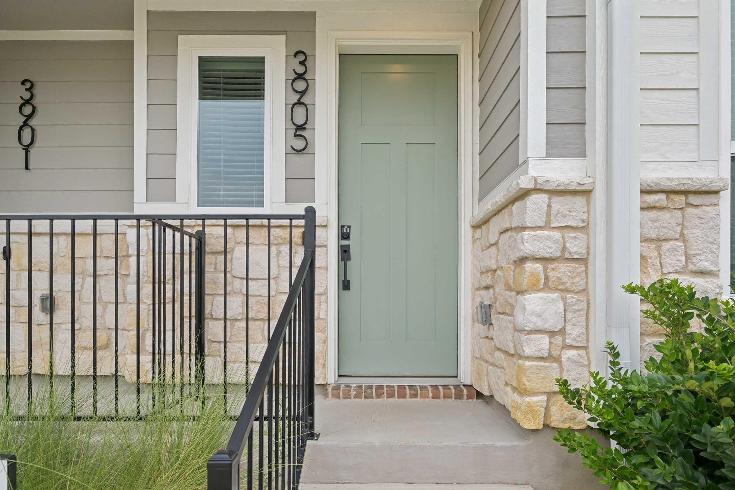 A light green front door with a black handle, set against a beige stone and gray siding exterior. The door is flanked by two house numbers: 3901 and 3905. A small front porch is visible with a wrought iron railing and some greenery at the base.