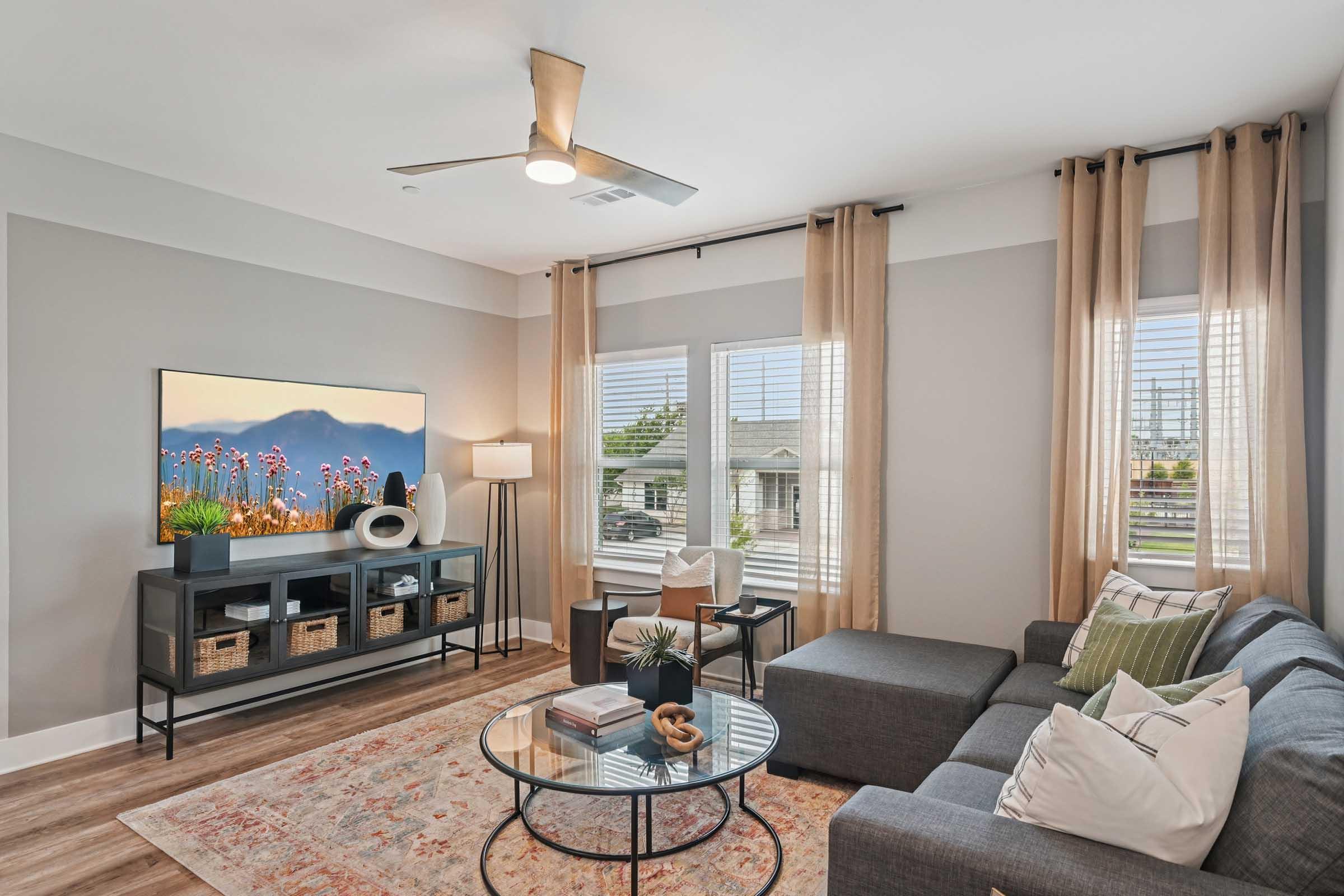 A modern living room featuring a gray sectional sofa, a decorative area rug, and a sleek TV mounted on the wall. Natural light streams through large windows with light curtains, complemented by a floor lamp and a stylish coffee table with decorative items.