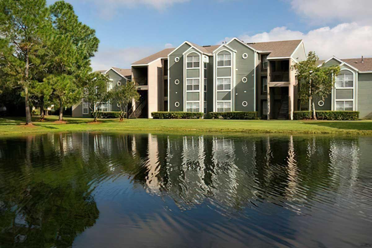 Two-story residential apartment buildings with green siding and large windows reflect in a calm pond.