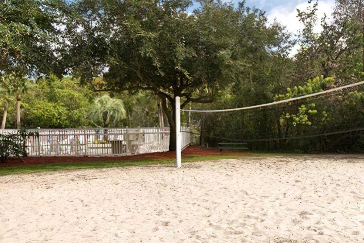 A sandy volleyball court surrounded by lush greenery, featuring a white net and a wooden bench in the background.