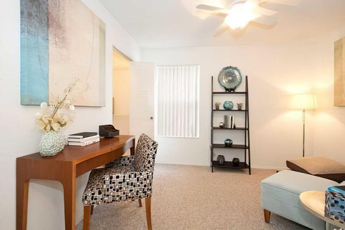 A cozy living space featuring a wooden desk with decorative items, a patterned chair, a bookshelf with art decor, and a soft blue ottoman.