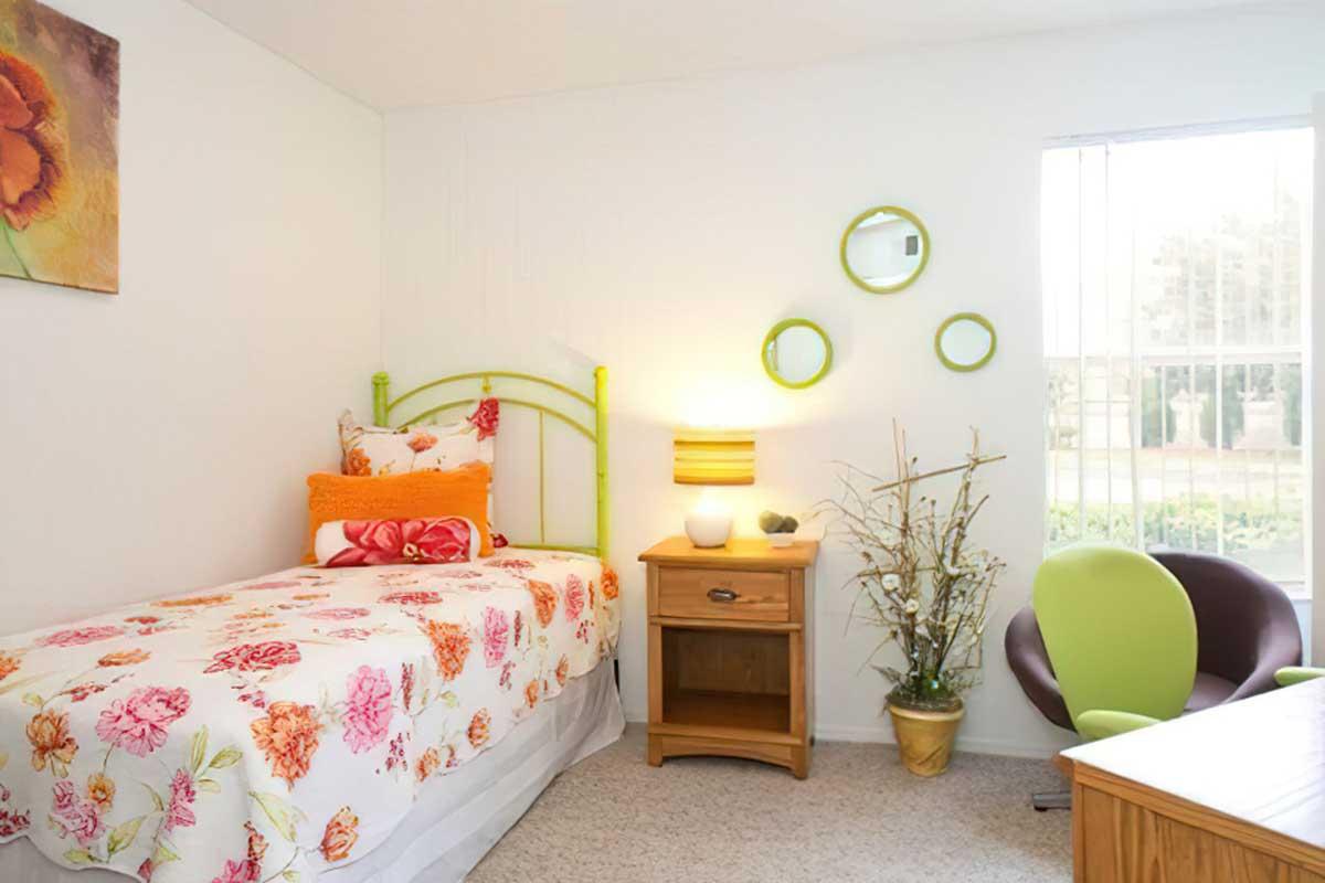 A brightly decorated bedroom featuring a green metal bed with floral bedding, a wooden nightstand with a lamp, a modern green chair, and wall decor including circular mirrors.