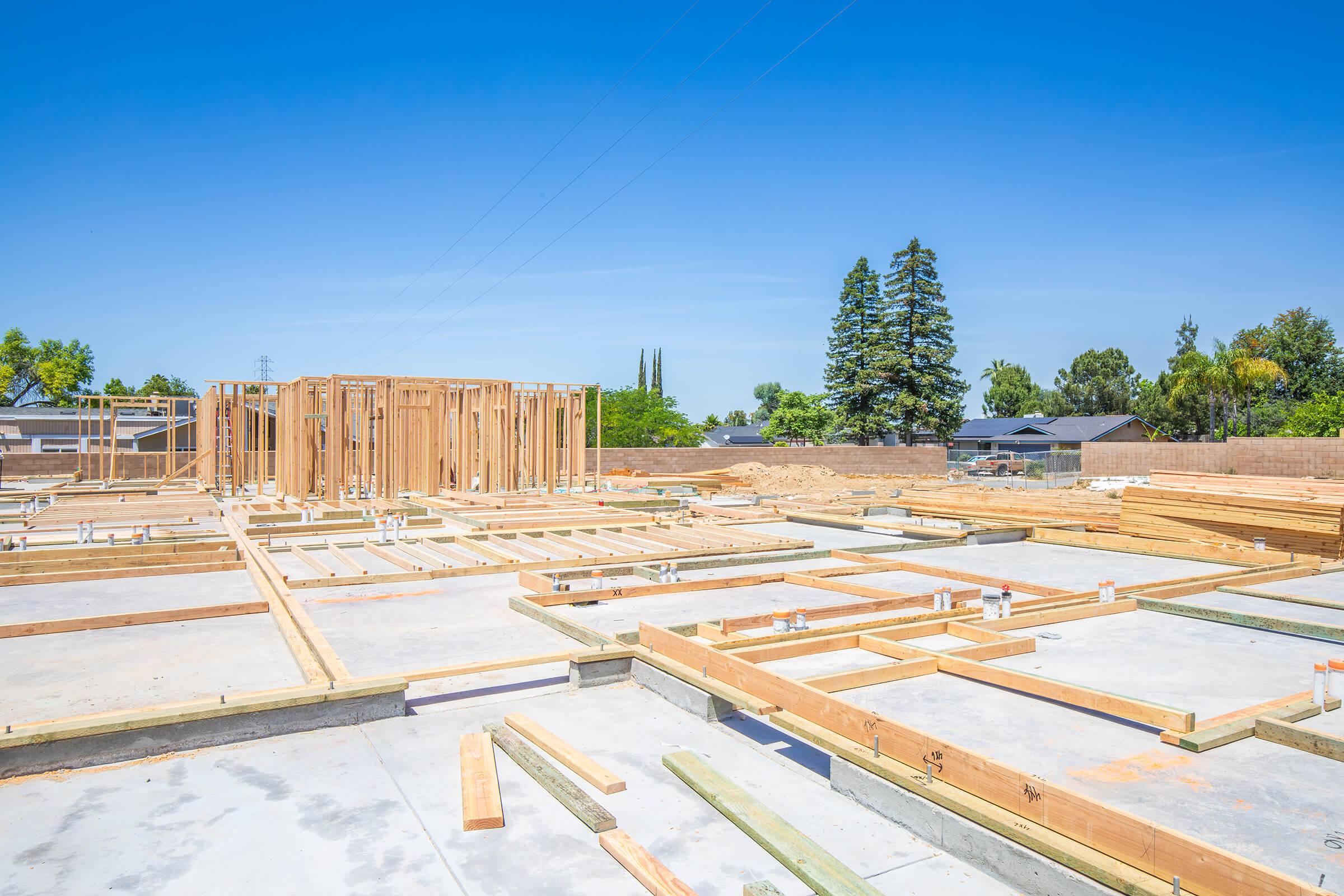 Construction site with a wooden frame structure in progress. The foundation is laid with concrete slabs, and wooden beams are arranged in a grid pattern. Blue sky overhead with a few trees and houses in the background, suggesting a residential area.