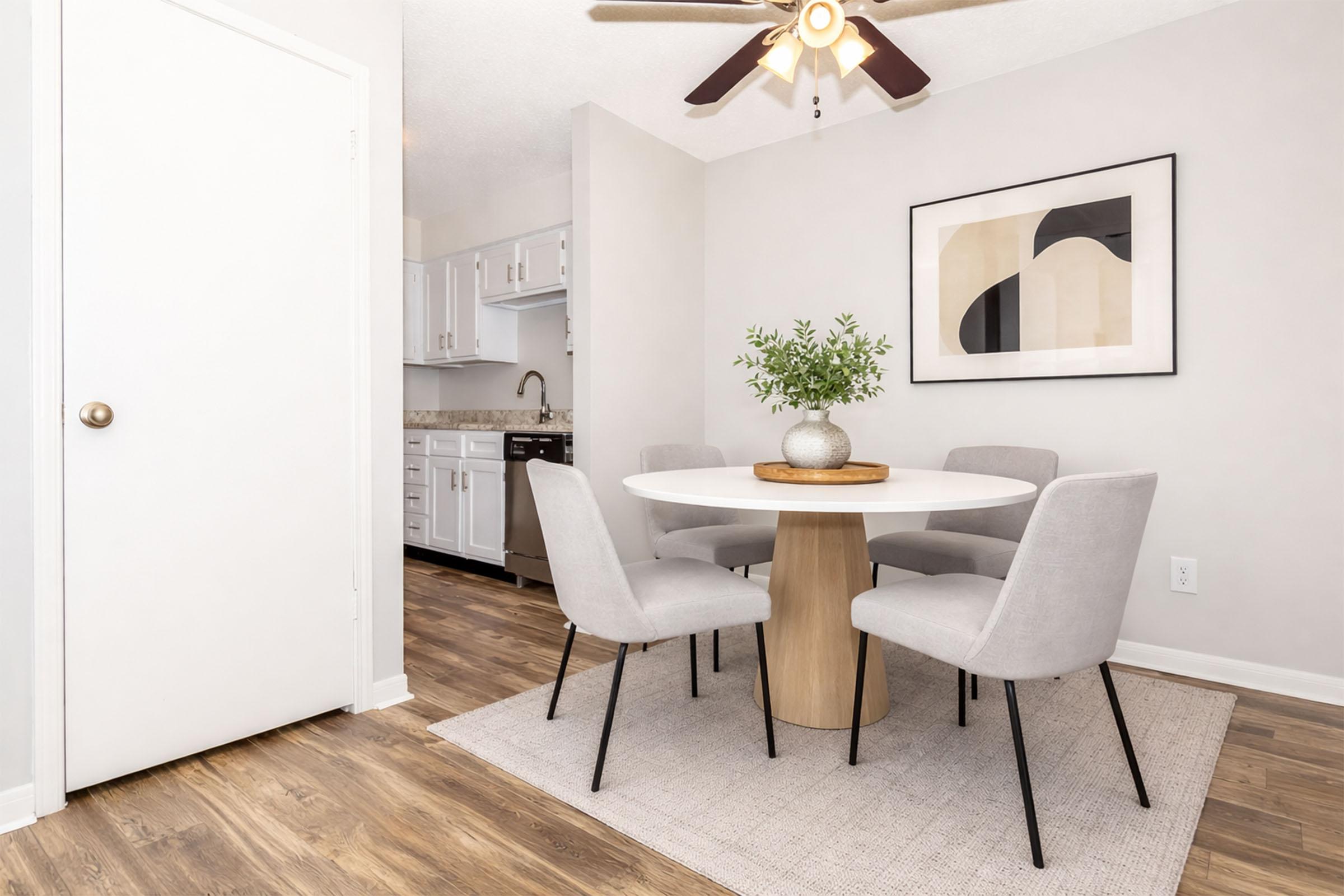 A modern dining area featuring a round white table surrounded by four light gray chairs. A potted plant sits at the center of the table. The walls are painted in neutral tones, and there is a ceiling fan above. In the background, a kitchen can be partially seen with white cabinetry.