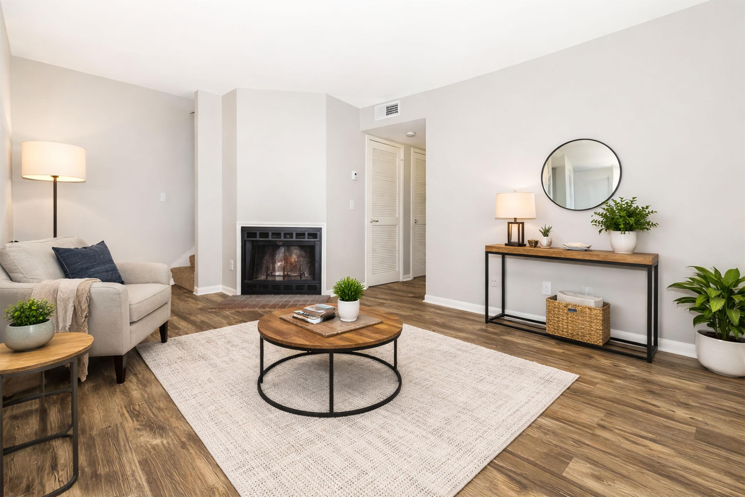 Cozy living room featuring a light-colored sofa, a round wooden coffee table, and a neutral area rug. A decorative mirror hangs on the wall, along with a stylish lamp and potted plants. A fireplace creates a warm ambiance, while soft lighting enhances the inviting atmosphere.