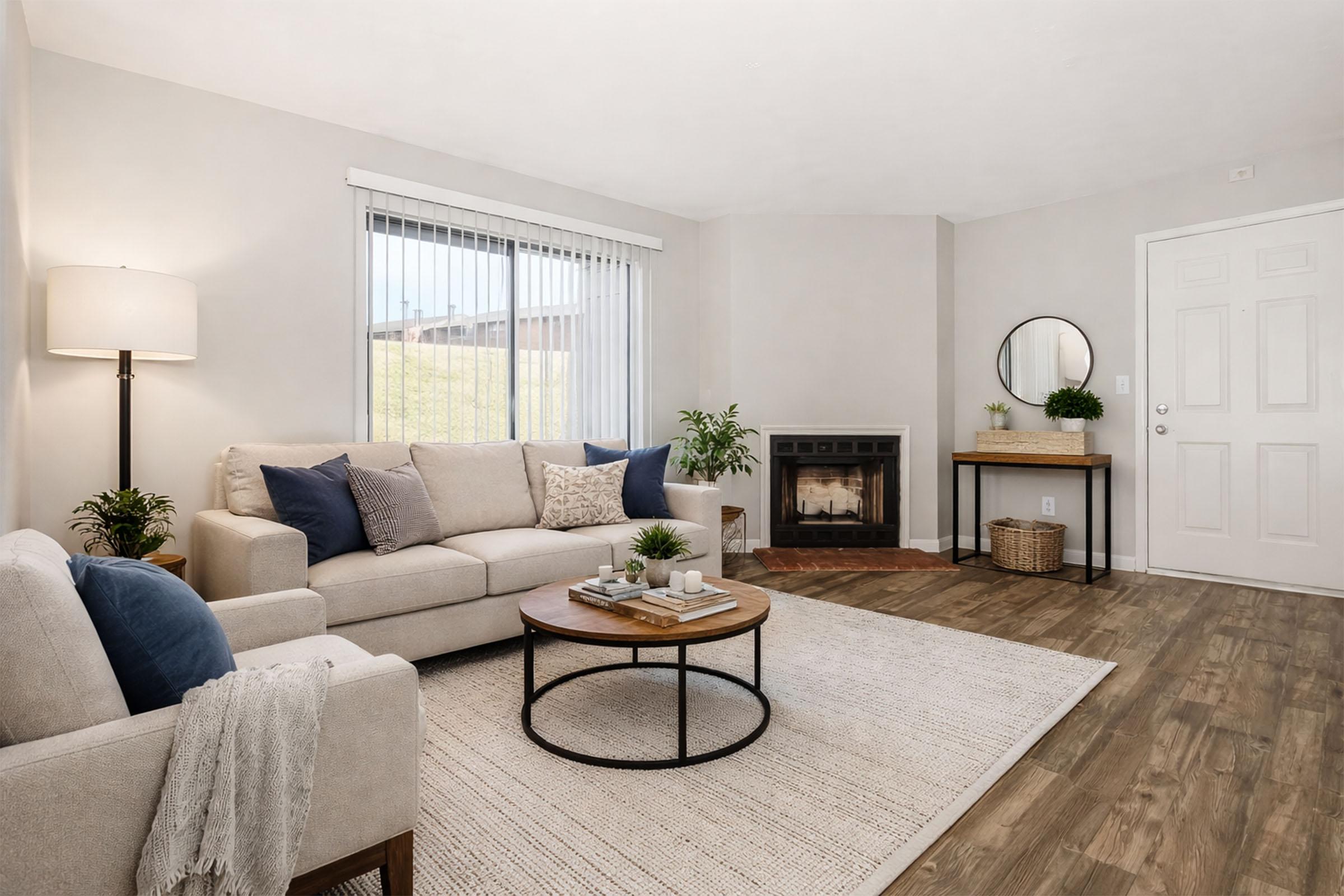 A cozy living room featuring a light-colored sofa with blue and white pillows, a round wooden coffee table, and a plush area rug. Natural light streams in through large windows with white blinds. A fireplace and a small table with a mirror complete the inviting space, surrounded by minimal decor and potted plants.