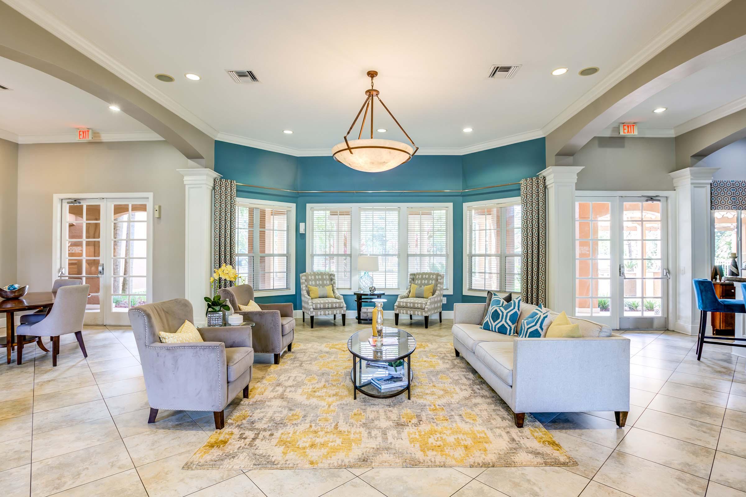 A bright and welcoming lobby area featuring a circular seating arrangement with plush armchairs and sofas, a stylish coffee table, and decorative accents. Large windows provide natural light, complemented by a modern chandelier overhead. The floor is tiled, adding to the elegant ambiance.