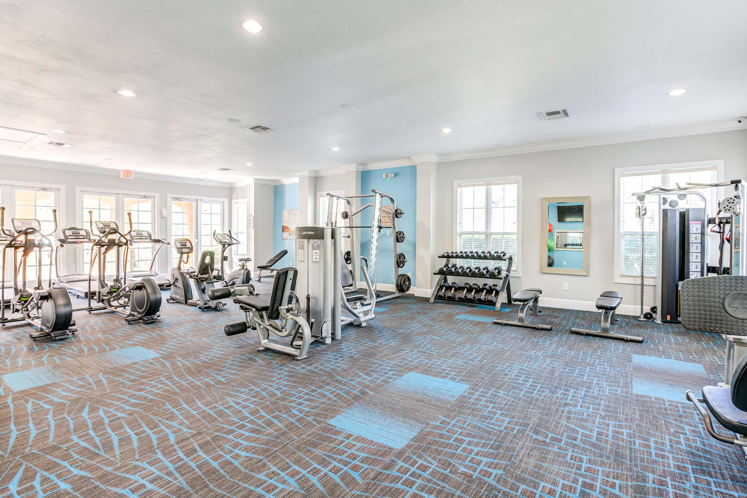 A modern gym featuring various workout equipment, including cardio machines like treadmills and ellipticals, weightlifting benches, and a rack of free weights. The bright space has large windows for natural light and a clean, inviting atmosphere with blue and gray carpeting.