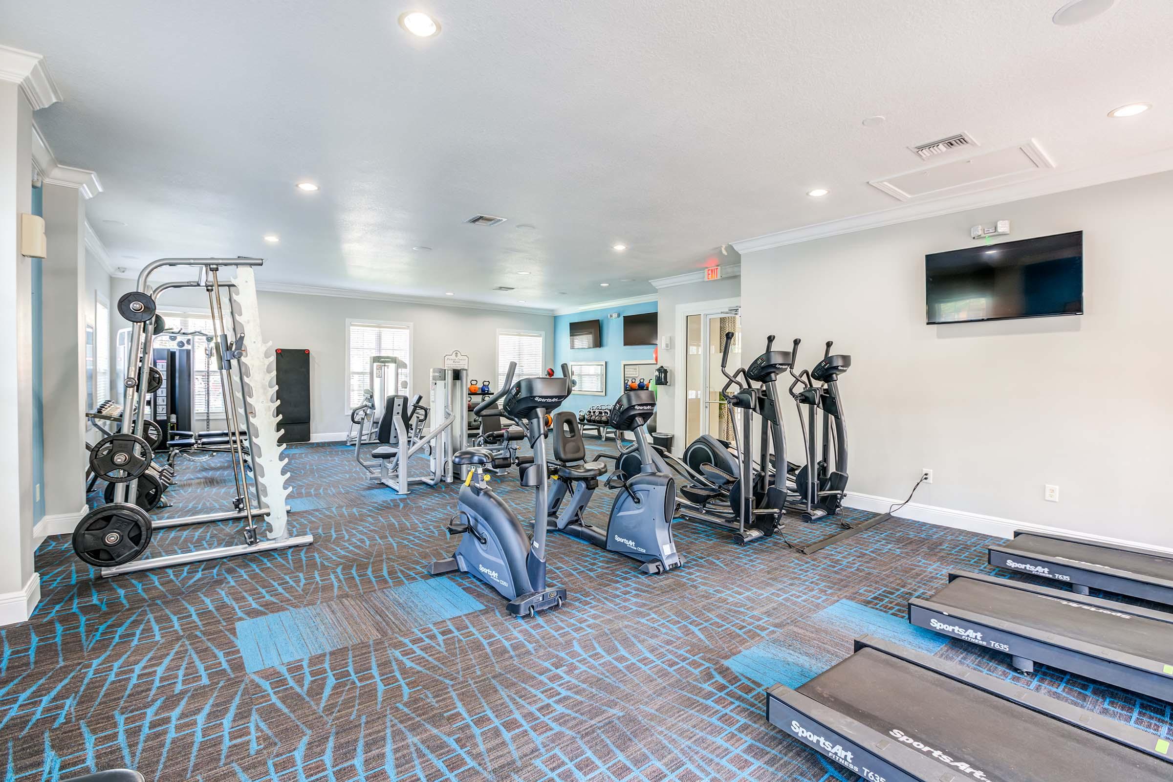 A modern gym interior featuring various exercise equipment, including treadmills, stationary bikes, weight machines, and free weights. The space is well-lit with a clean design and has mirrored walls for an open feel.