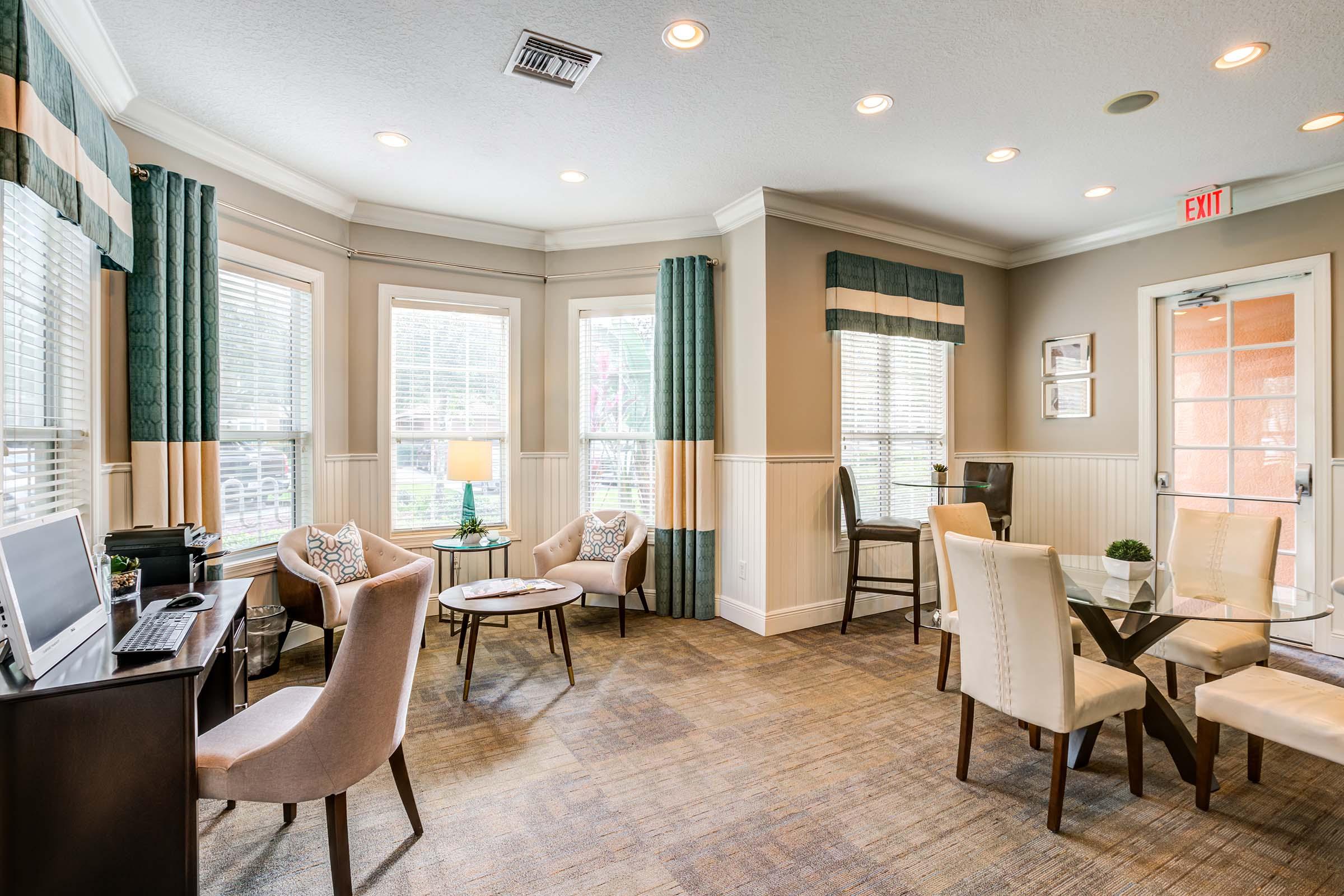 A cozy sitting area with large windows, soft lighting, and a mix of chairs and tables. There’s a desk with a computer, potted plants, and decorative cushions on the chairs. The walls are painted in neutral tones, and there's a door leading outside. Overall, a welcoming and bright atmosphere.