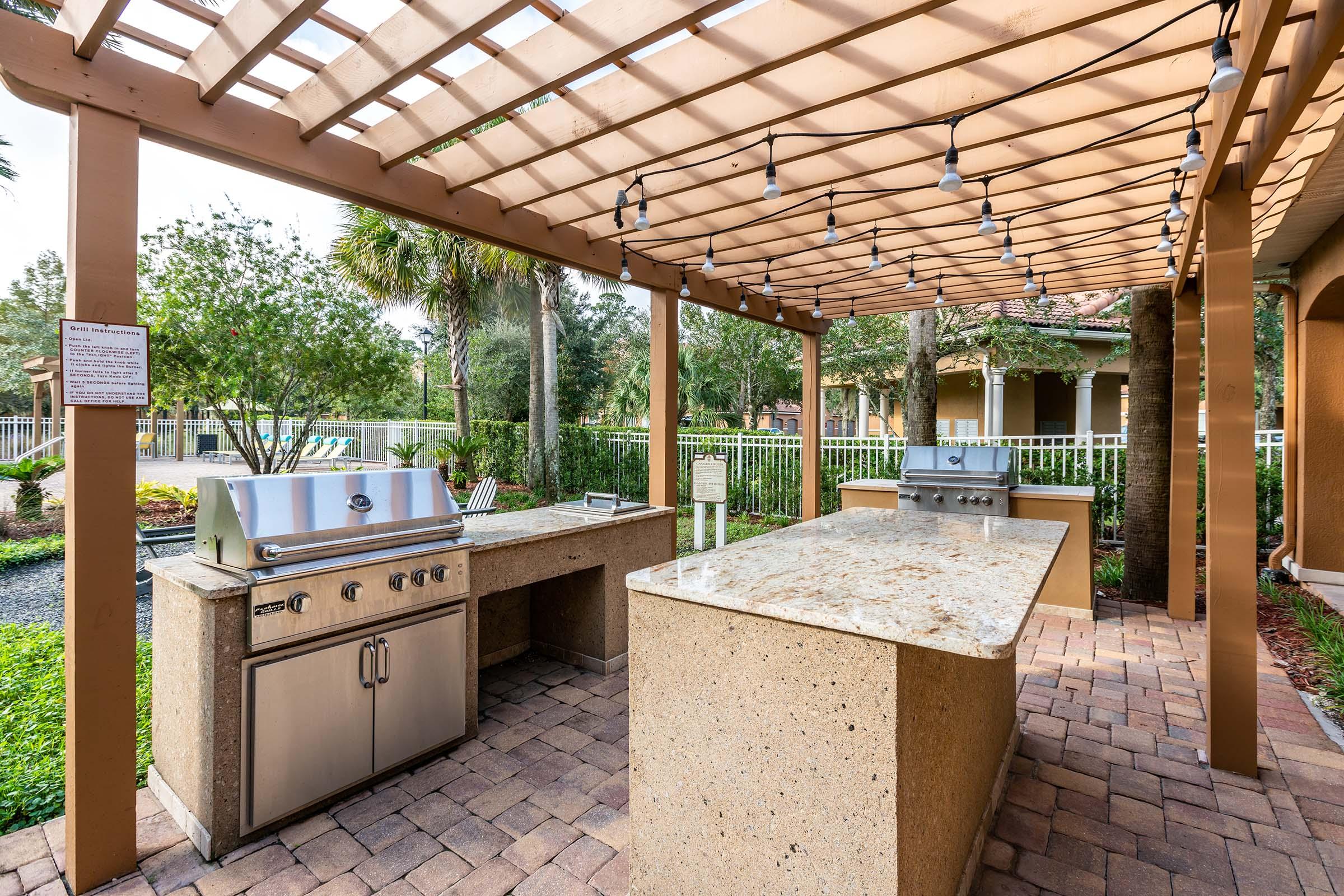 An outdoor kitchen featuring a pergola with string lights, a built-in grill, and a large countertop. The area is surrounded by lush greenery and landscaping, providing a welcoming space for outdoor cooking and dining.