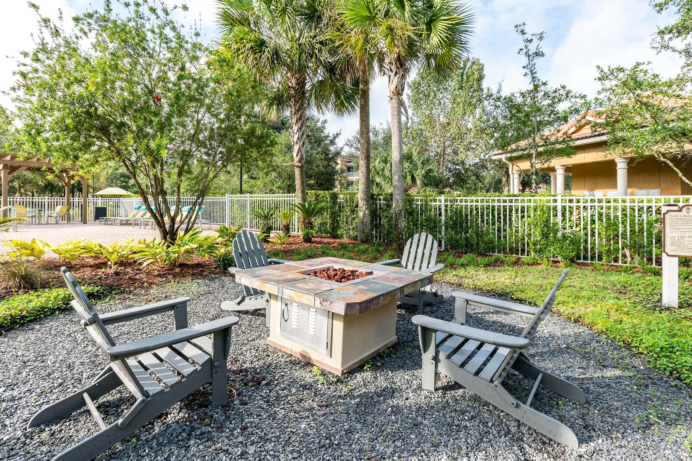 A cozy outdoor seating area featuring a stone fire pit surrounded by four wooden Adirondack chairs. Lush greenery and palm trees provide a tropical backdrop, with a swimming pool visible in the distance. Clear blue skies and well-maintained landscaping create a relaxing atmosphere.