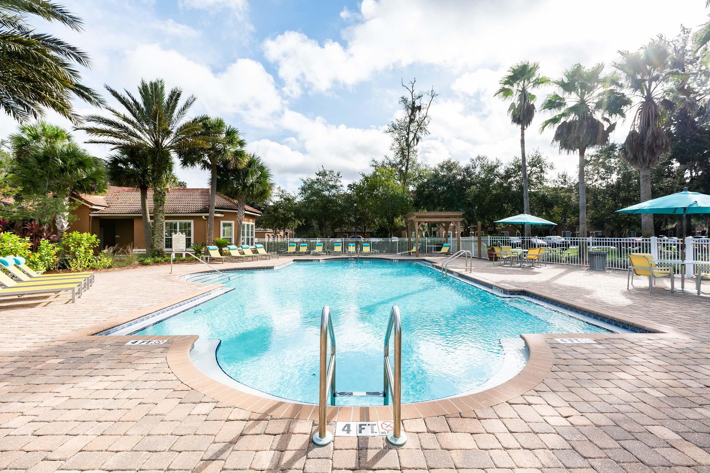 A bright outdoor pool area featuring clear blue water, surrounded by palm trees and lounge chairs. A cheerful atmosphere with a nearby cabana and umbrellas. The pool has a depth marker at 4 feet and is framed by a paved deck, inviting for relaxation and leisure under a partly cloudy sky.
