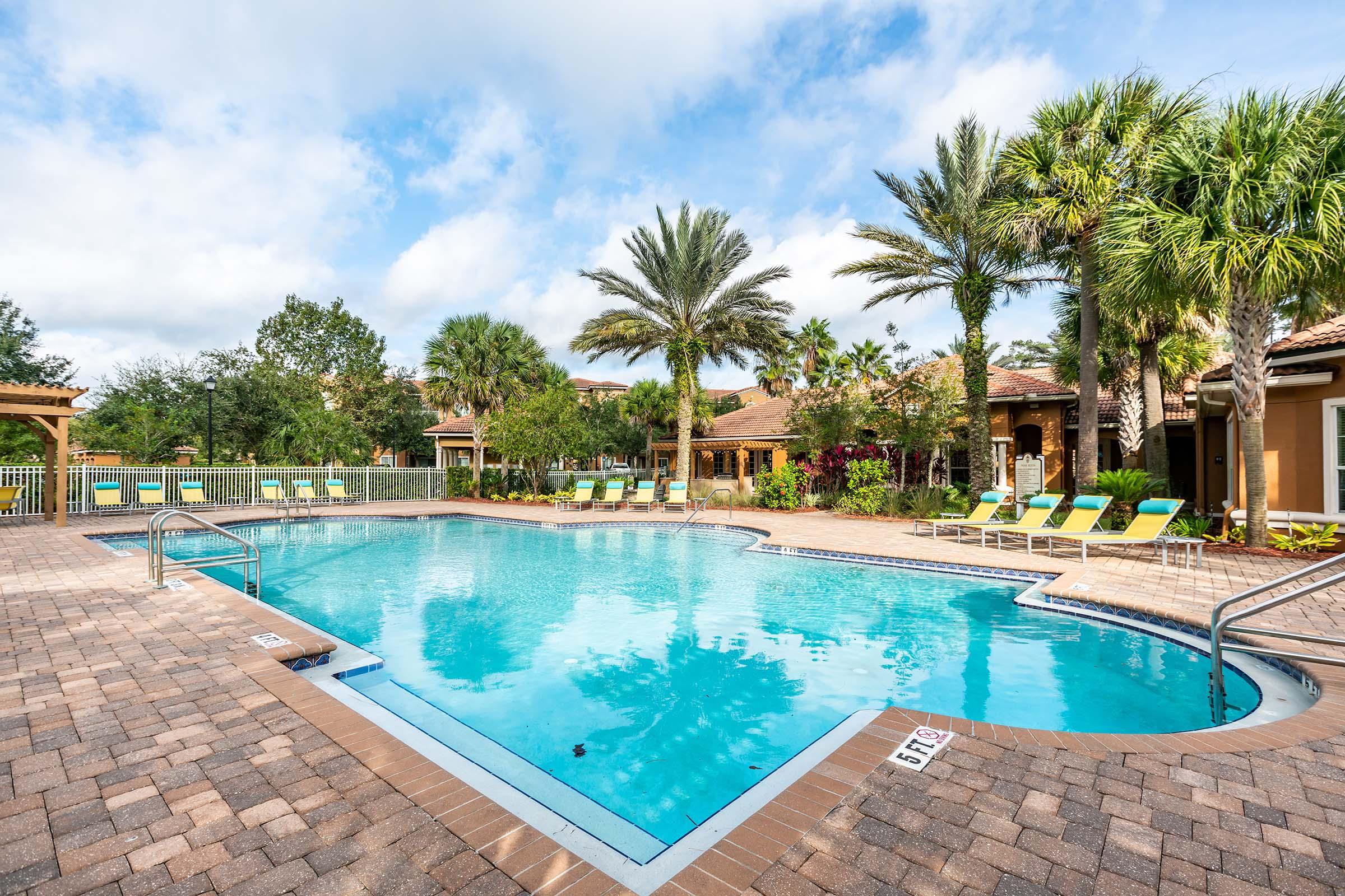 A tranquil outdoor swimming pool surrounded by palm trees and lounge chairs. The pool area is bordered by brick pavement, with a pergola in the background and a lush, green landscape reflected in the clear water under a partly cloudy sky.