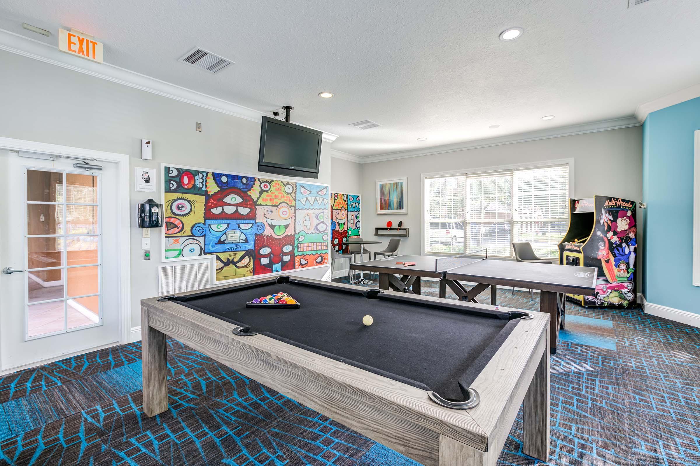 Interior of a recreational room featuring a billiard table with a pool game in progress. A colorful mural decorates the wall, and there are arcade games and a seating area. Large windows allow natural light, and there's a TV mounted on the wall.