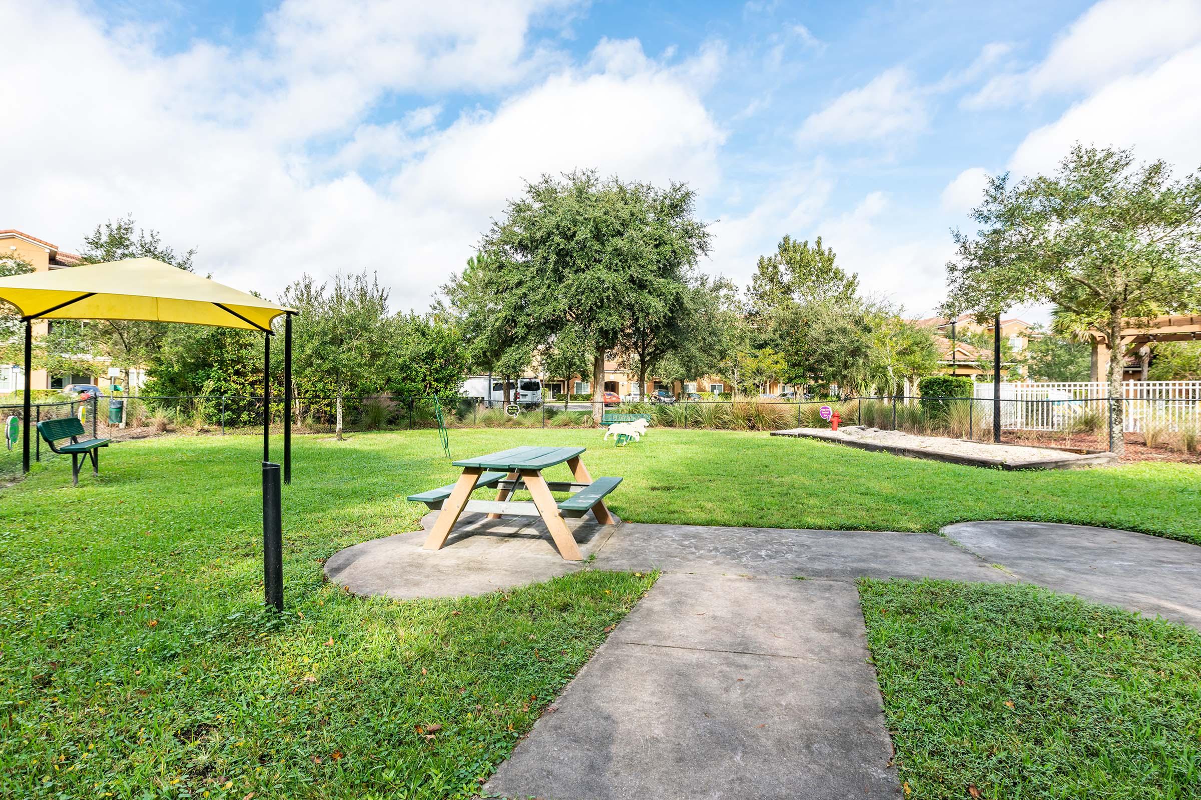 A tranquil park scene featuring a green picnic table under a yellow canopy, surrounded by grassy areas and trees. A pathway meanders through the park, leading to a fenced area in the background. The sky is partly cloudy, adding a serene ambiance to the outdoor space.