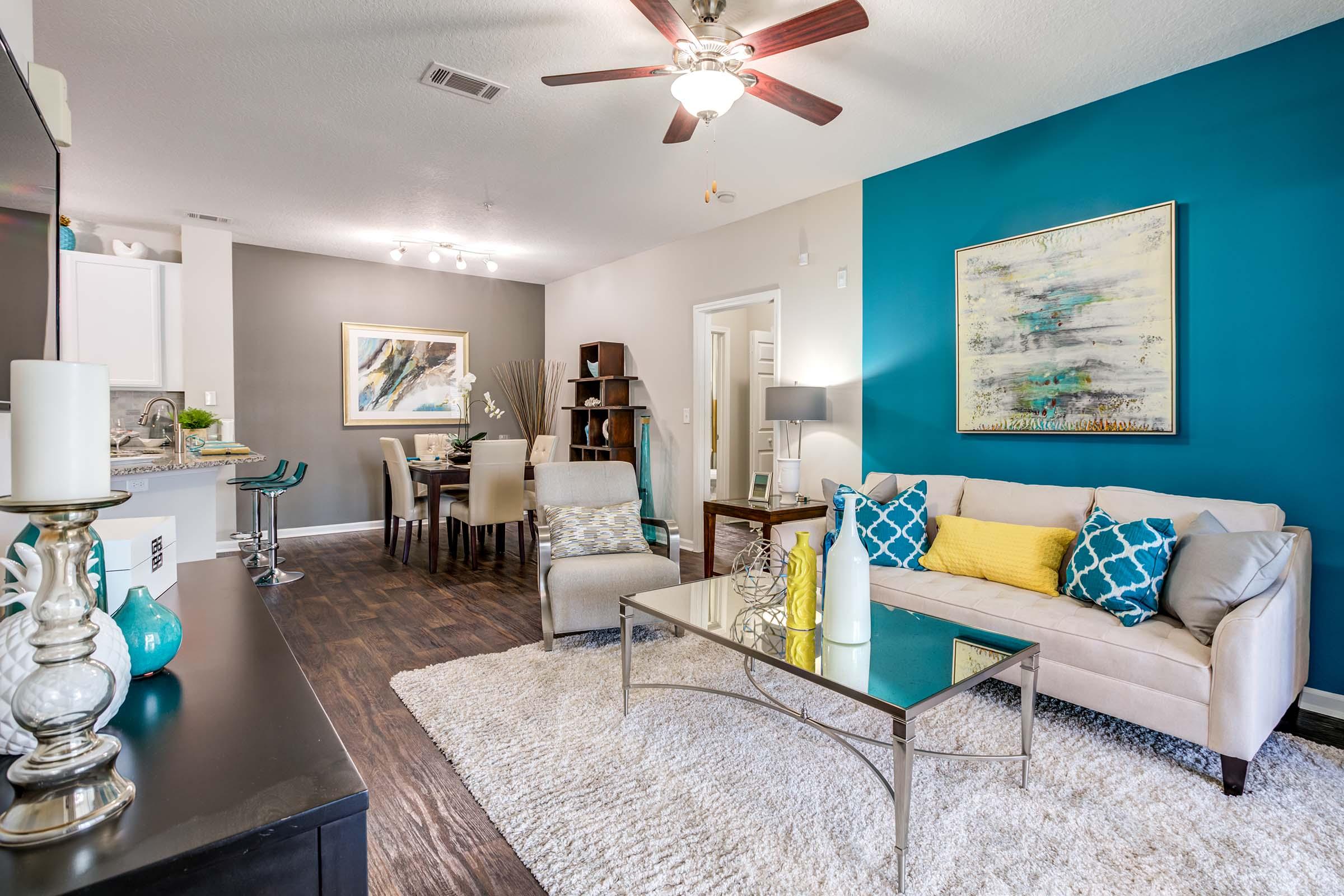 A modern living room featuring a beige sofa with blue and yellow pillows, a glass coffee table on a large area rug, and stylish decor. A teal accent wall complements the neutral tones, and there is a dining area visible in the background with a round table and chairs. Large windows allow for natural light.