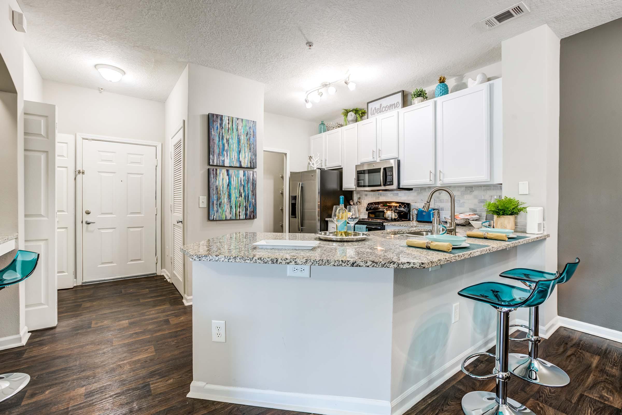 Modern kitchen featuring a granite countertop island with three blue bar stools, stainless steel appliances, and white cabinetry. The space is well-lit with overhead lights and has decorative items on the shelves. A door leads to the entry area, and there's a textured wall in a darker shade.