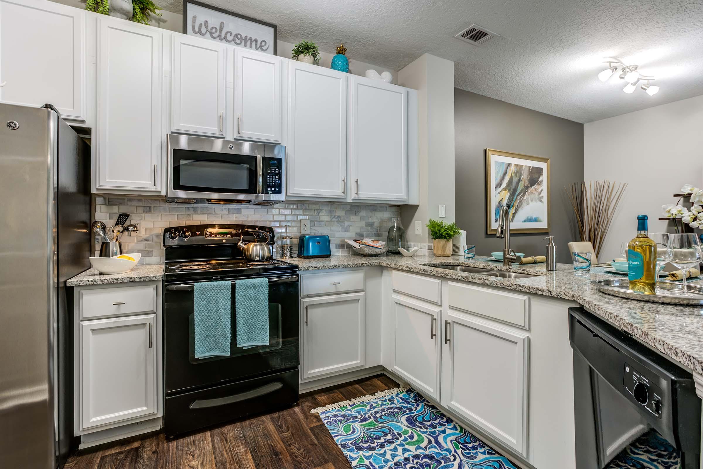 Modern kitchen featuring white cabinetry, a stainless steel refrigerator, and a black stove. The countertop is adorned with granite, and a decorative rug in blue and green adds a pop of color. Bright lighting and tasteful decor, including a plant and art piece, enhance the inviting atmosphere.