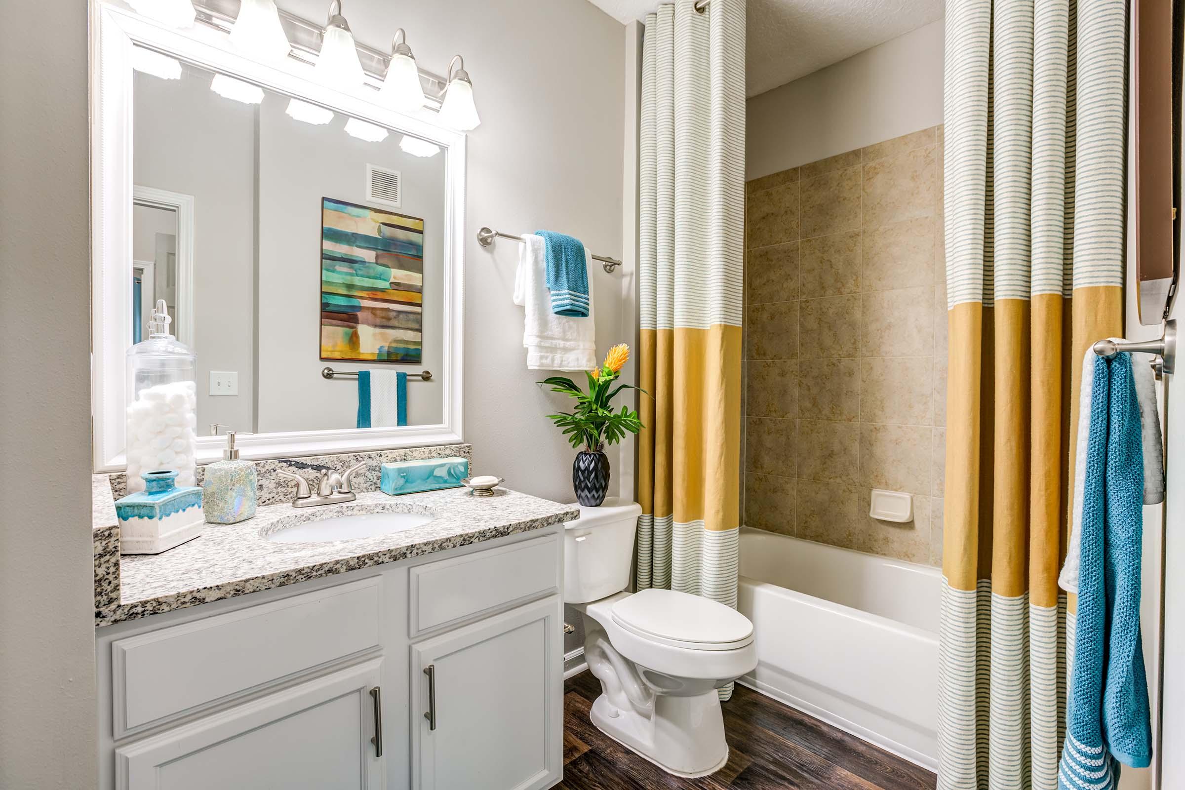 A modern bathroom featuring a white vanity with a granite countertop, a large mirror above it, and decorative items. The bathtub is enclosed by a striped shower curtain in shades of yellow and beige. Fresh flowers are placed in a vase on the counter, adding a touch of color to the neutral decor.